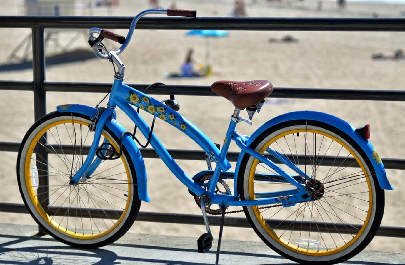Beach Cruiser 桌布 壁紙 — 一輛亮藍色沙灘巡洋艦自行車，配有黃色輪圈和向日葵貼花，靠在俯瞰陽光沙灘的欄杆上。