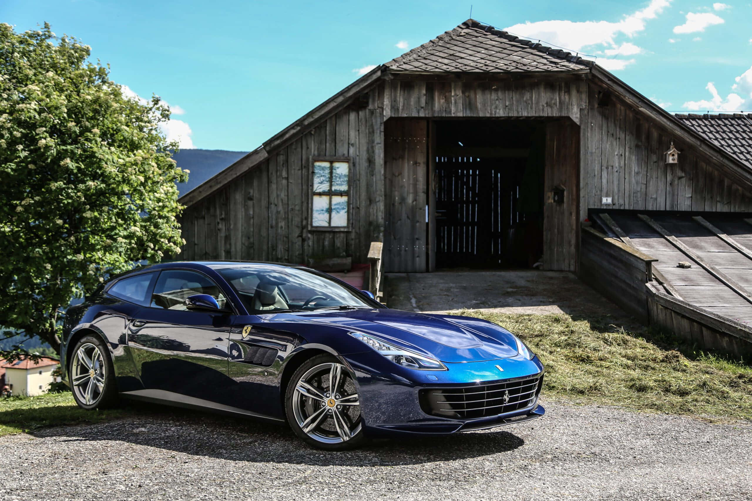 Ferrari Gtc4lusso 桌布 壁紙 — 一輛亮藍色法拉利停在碎石路上，前面是質樸的木製穀倉，上方是明亮的藍天。