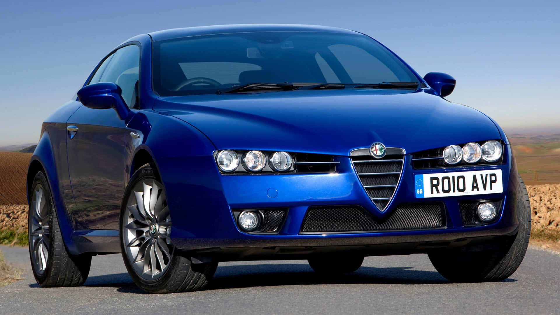 Alfa Romeo Brera 桌布 壁紙 — 一輛亮藍色的愛快羅密歐跑車停泊在鋪砌的道路上，乾燥的山丘在清澈的藍天下。