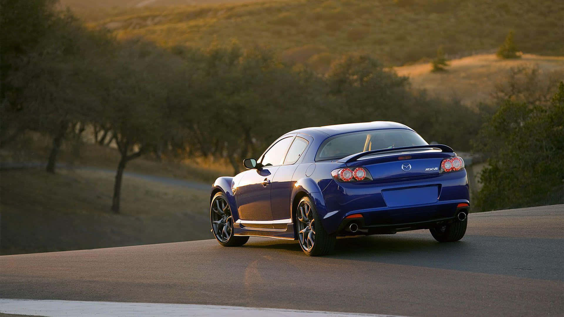 Mazda Rx-8 桌布 壁紙 — 一輛亮藍色跑車在日落時分沿著蜿蜒的道路穿過金黃色的山丘行駛。