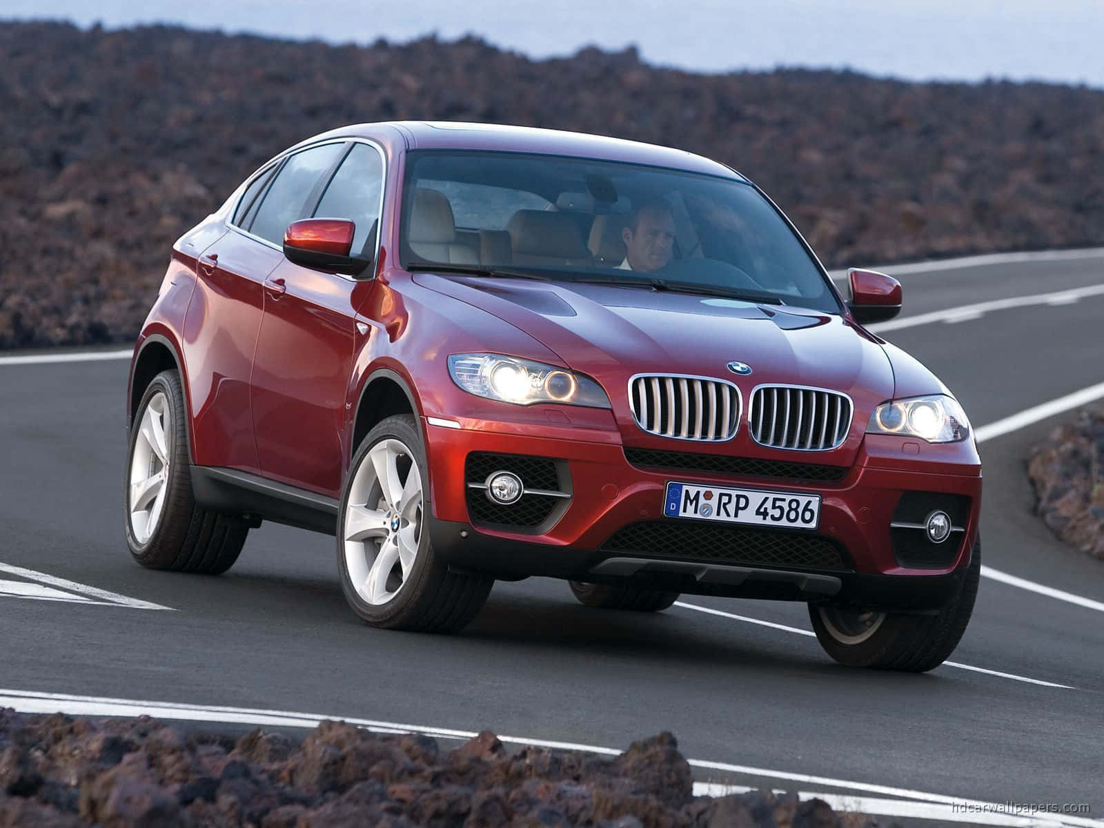 Bmw X6 桌布 壁紙 — 一輛亮面紅色BMW X6在晴朗天空下，沿著崎嶇火山地形兩旁蜿蜒的道路行駛。