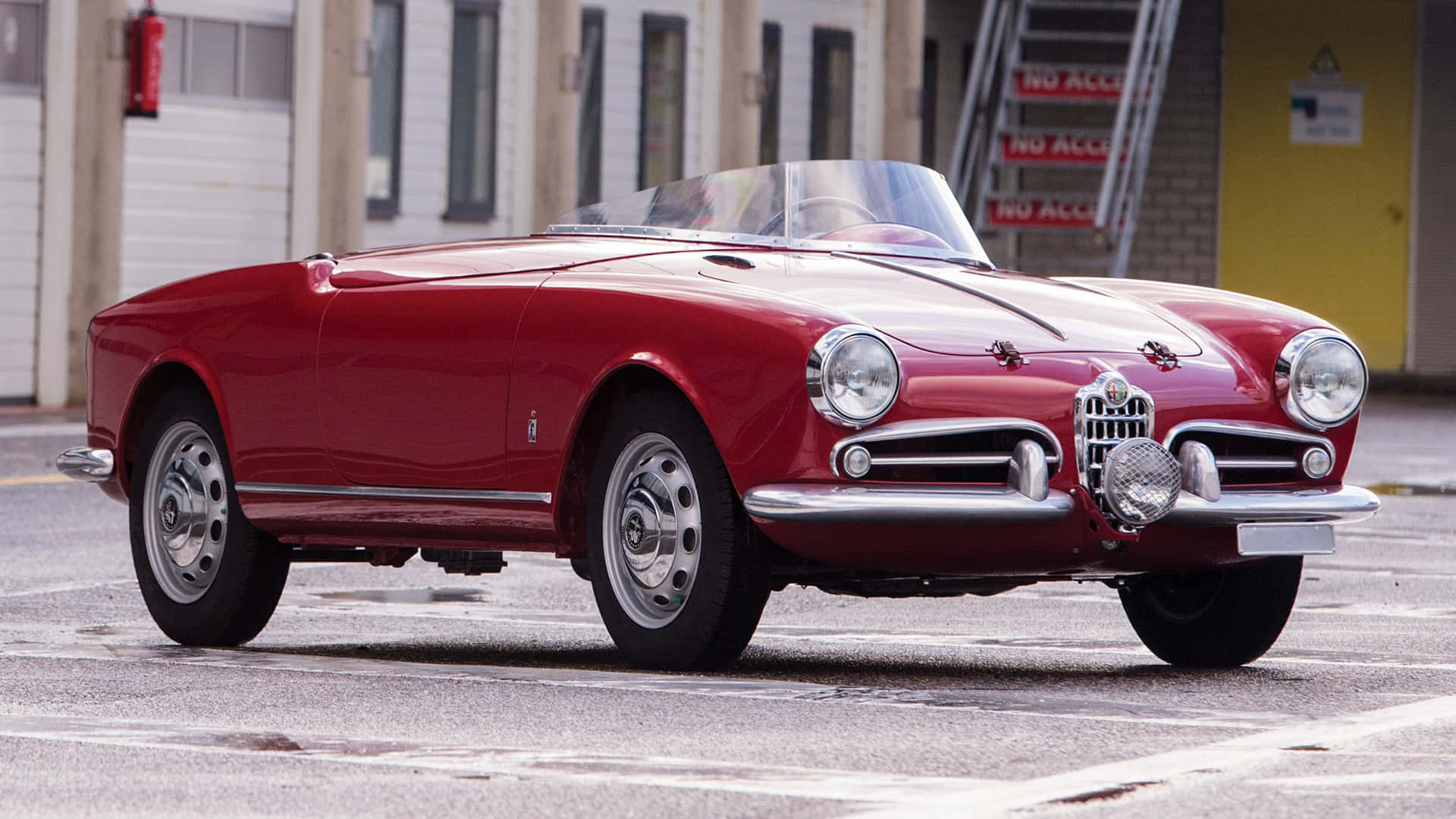 Alfa Romeo Spider 桌布 壁紙 — 一輛亮面紅色復古愛快羅密歐Spider開篷車停泊在濕瀝青地上，背景有工業建築。