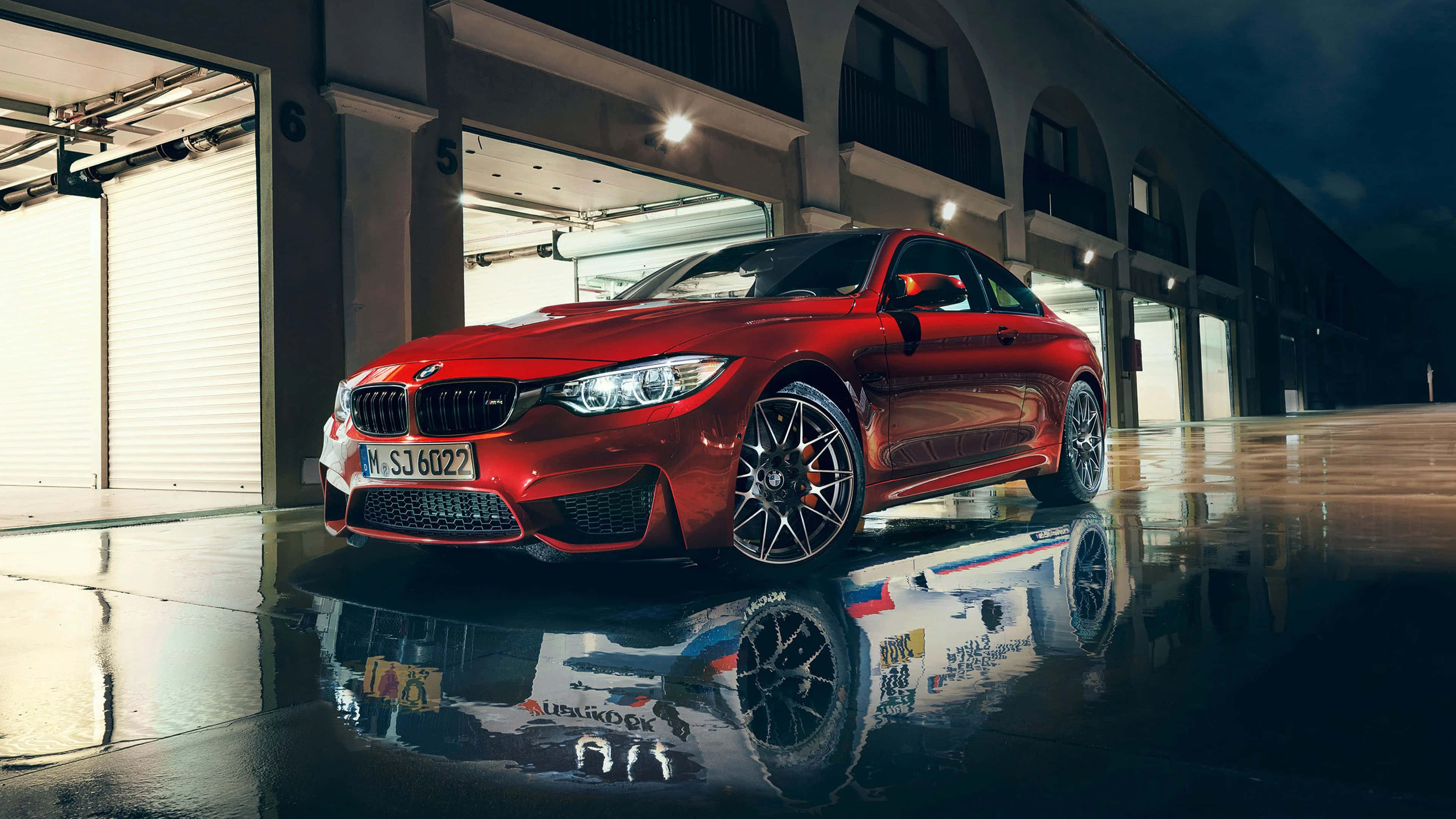 4K BMW 壁紙 桌布 壁紙 — 一輛亮麗的紅色BMW M4轎跑車在夜燈下閃耀，其倒影在工業車庫下的濕漉漉路面上閃爍。