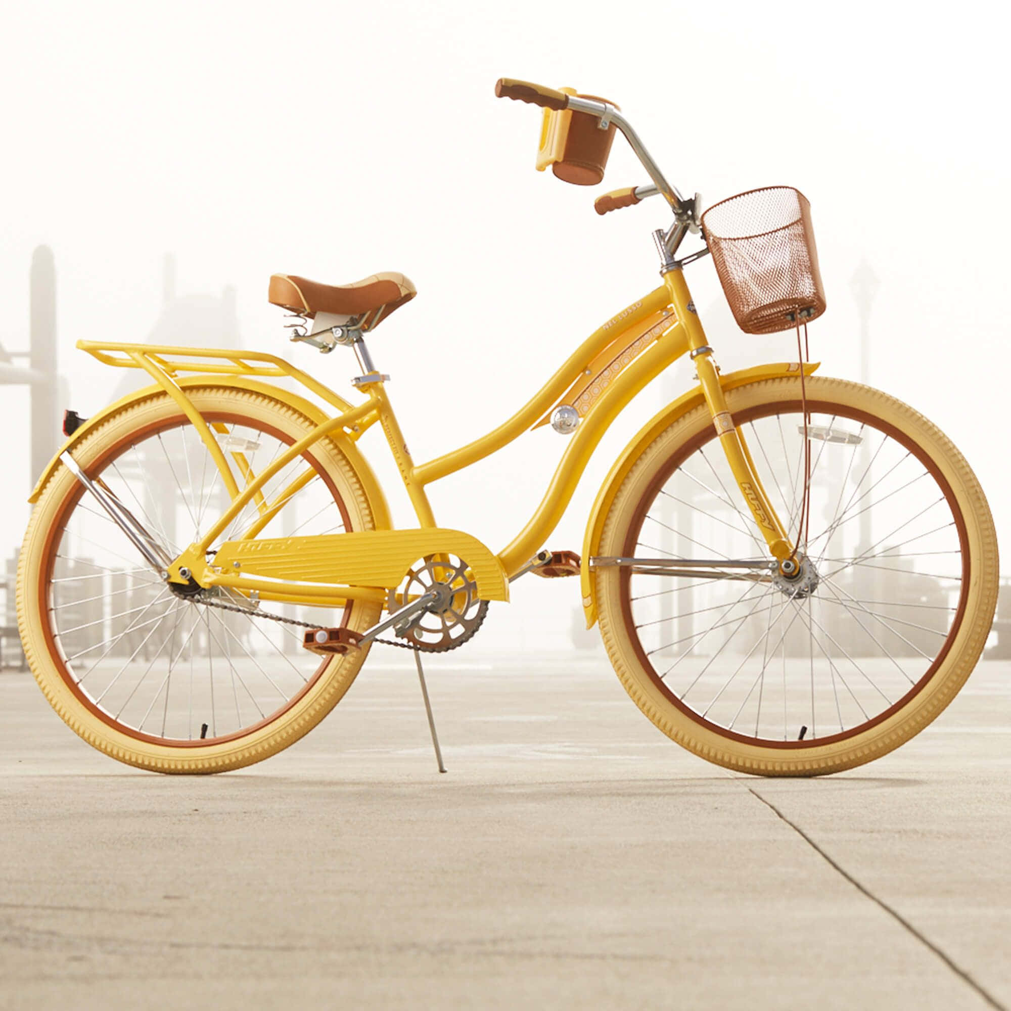 Beach Cruiser 桌布 壁紙 — 一輛亮黃色復古單車，配有棕色籃子和座椅，立在混凝土表面上，背景是朦朧的城市景觀。