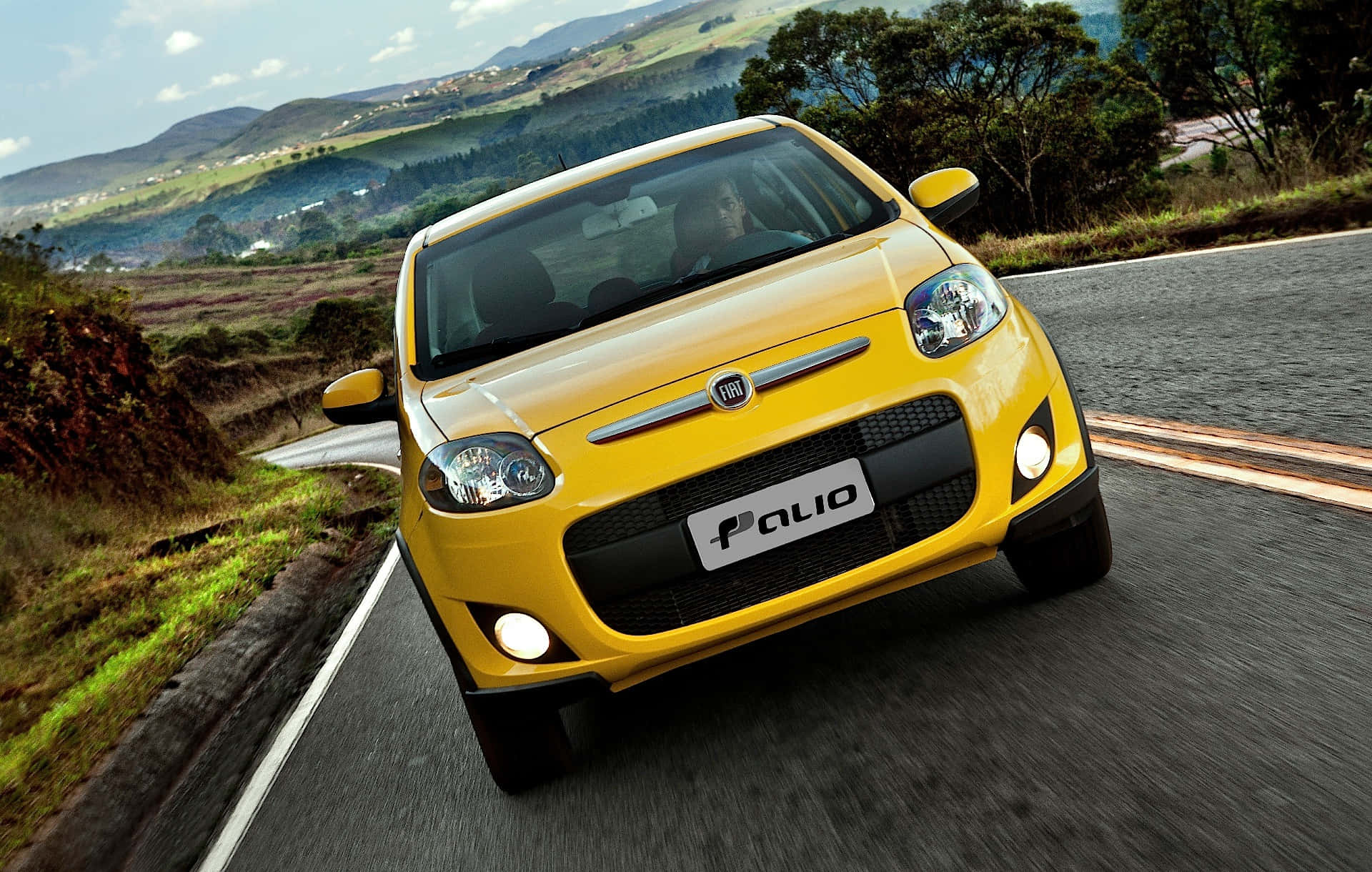 Fiat Palio 桌布 壁紙 — 一輛亮黃色的Fiat Palio在蜿蜒的山路上行駛，周圍是茂密的綠色山丘和樹木，天空部分多雲。