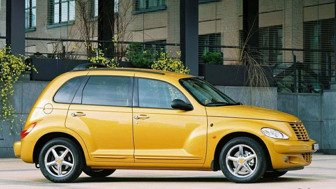 Chrysler Pt Cruiser 桌布 壁紙 — 一輛亮黃色的PT Cruiser停泊在現代玻璃建築旁的城市行人路上，旁邊有開花的灌木叢。