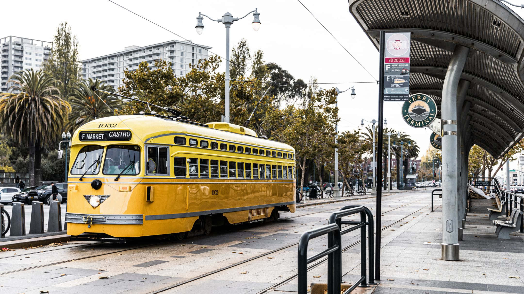 三藩市 桌布 壁紙 — 一輛亮黃色的復古纜車，標有 F Market Castro，在陰天下滑過軌道，經過棕櫚樹和現代建築。