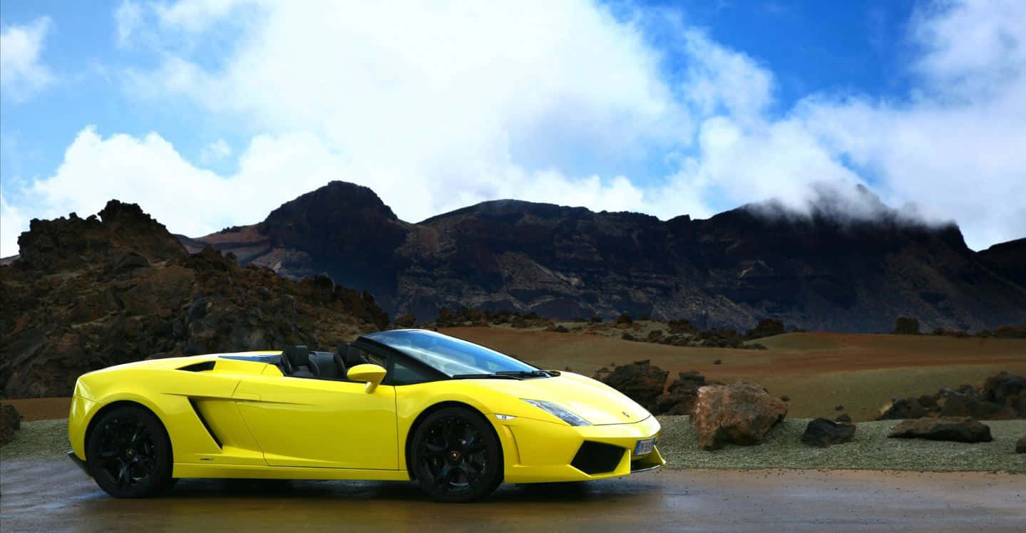 Lamborghini Gallardo 桌布 壁紙 — 一輛亮黃色的林寶堅尼Gallardo Spyder停泊在濕滑的道路上，背景是崎嶇的火山山脈，天空部分多雲。