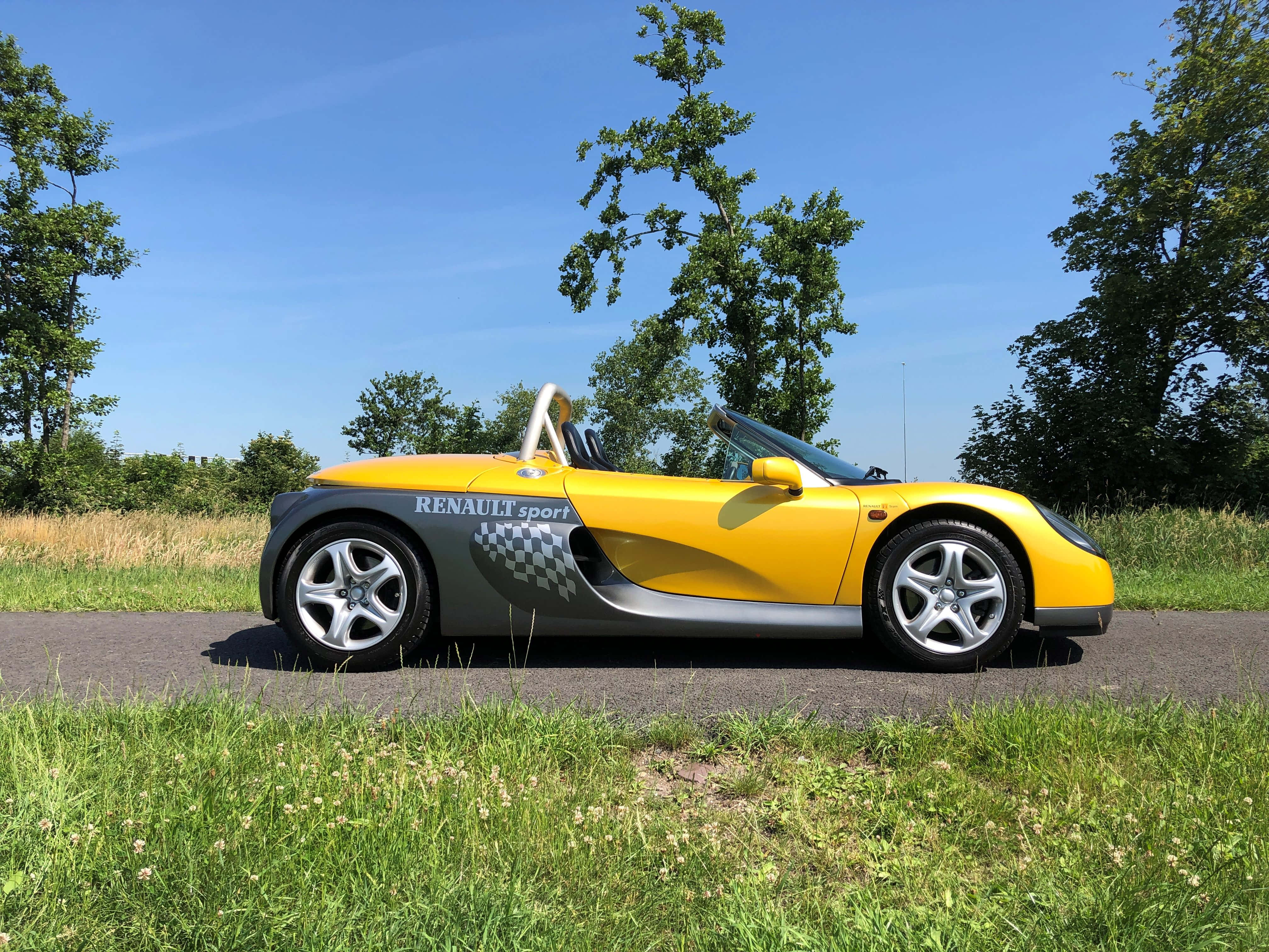 Renault Sport Spider 桌布 壁紙 — 一輛亮黃色的雷諾運動蜘蛛敞篷車停在鄉間小路上，天空湛藍，綠樹成蔭，草地翠綠。