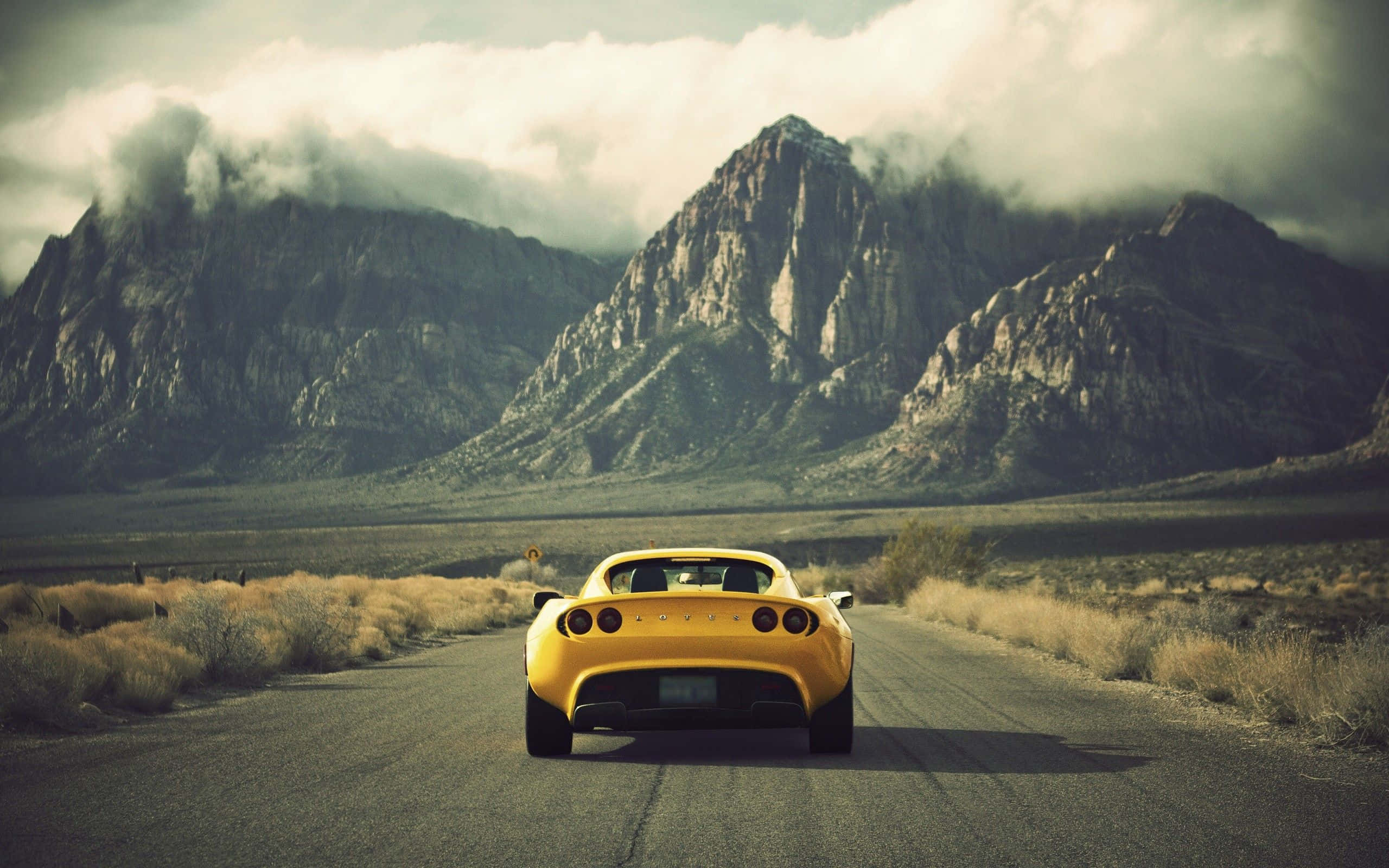 Yellow Car 桌布 壁紙 — 一輛亮黃色跑車在沙漠公路上行駛，駛向雲霧繚繞的高聳岩石山脈。