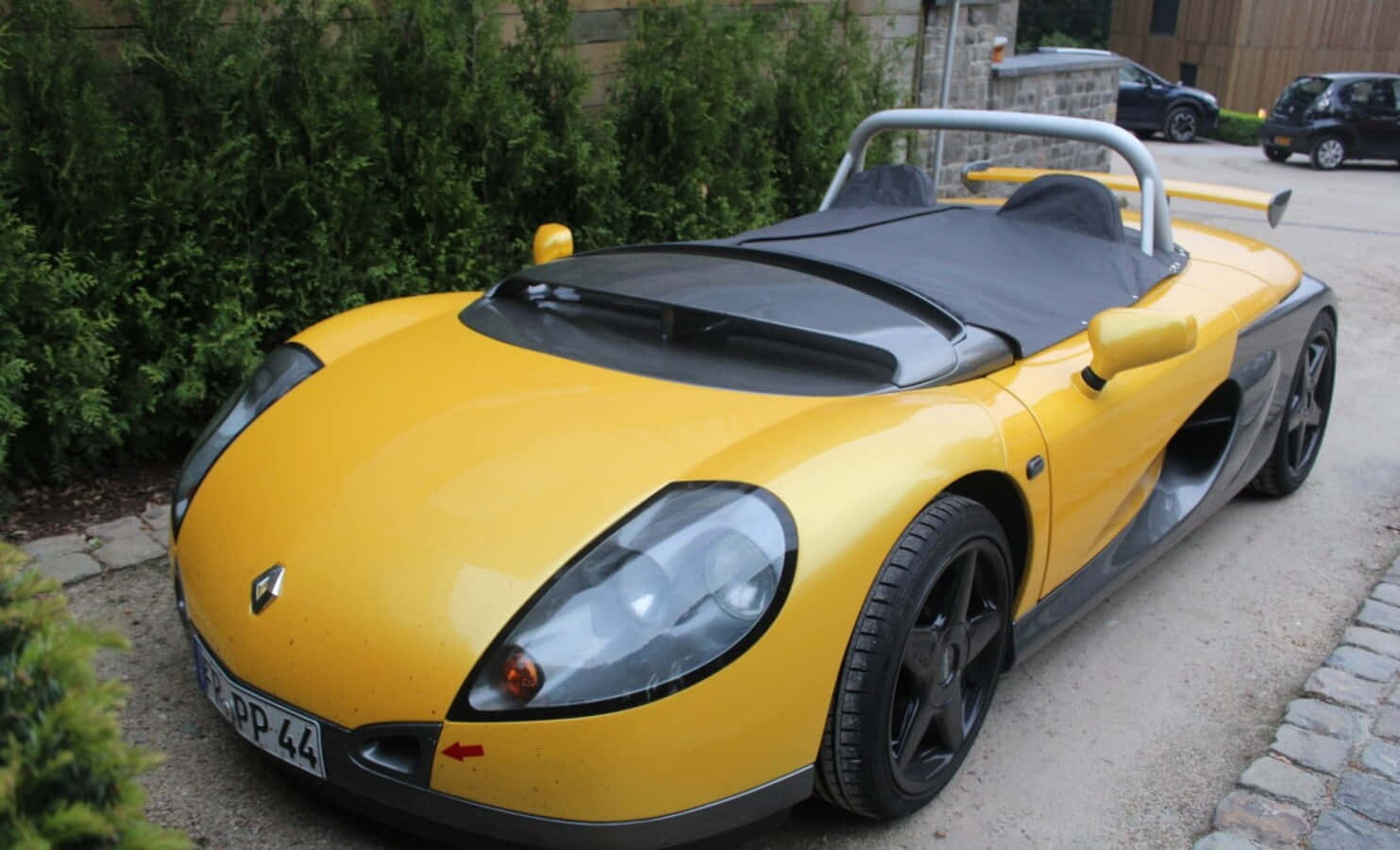 Renault Sport Spider 桌布 壁紙 — 一輛亮黃色雷諾Spider跑車，帶黑色飾邊，停在鵝卵石車道上，旁邊是綠色籬笆。