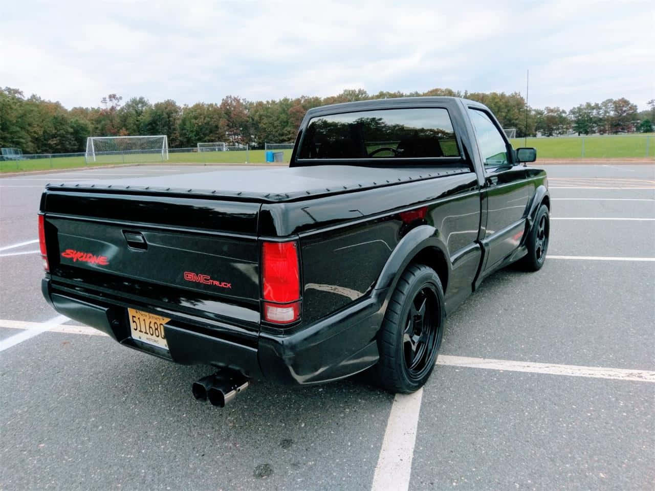 Gmc Syclone 桌布 壁紙 — 一輛亮黑色GMC Sonoma皮卡車停在瀝青停車場，背景有樹木和足球場。