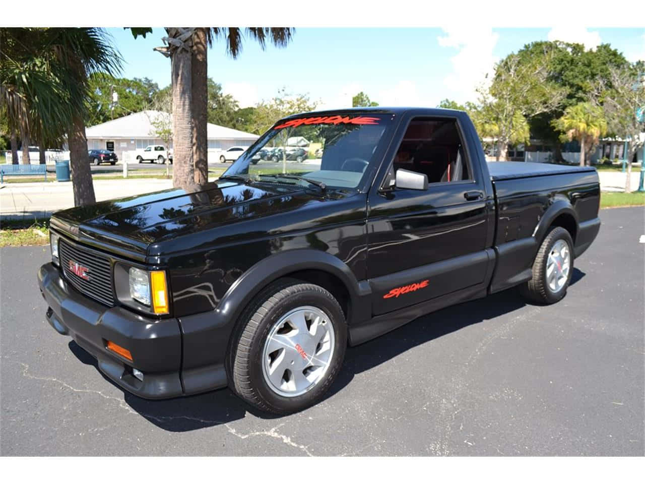 Gmc Syclone 桌布 壁紙 — 一輛亮黑色GMC Syclone皮卡車停在瀝青路面上，在明亮的日光下，背景有棕櫚樹和房屋。