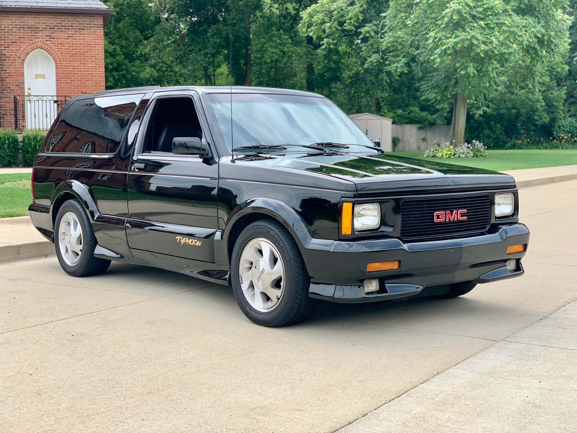 Gmc Typhoon 桌布 壁紙 — 一輛亮黑色的GMC Typhoon SUV停放在混凝土車道上，背景是磚房和綠樹。