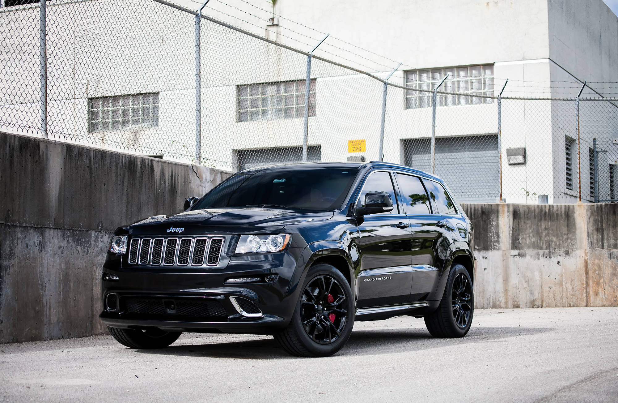 Jeep Grand Cherokee SRT 壁紙 — 一輛亮黑色的Jeep Grand Cherokee SRT停在鏈條圍欄和混凝土牆前，在明亮的日光下。