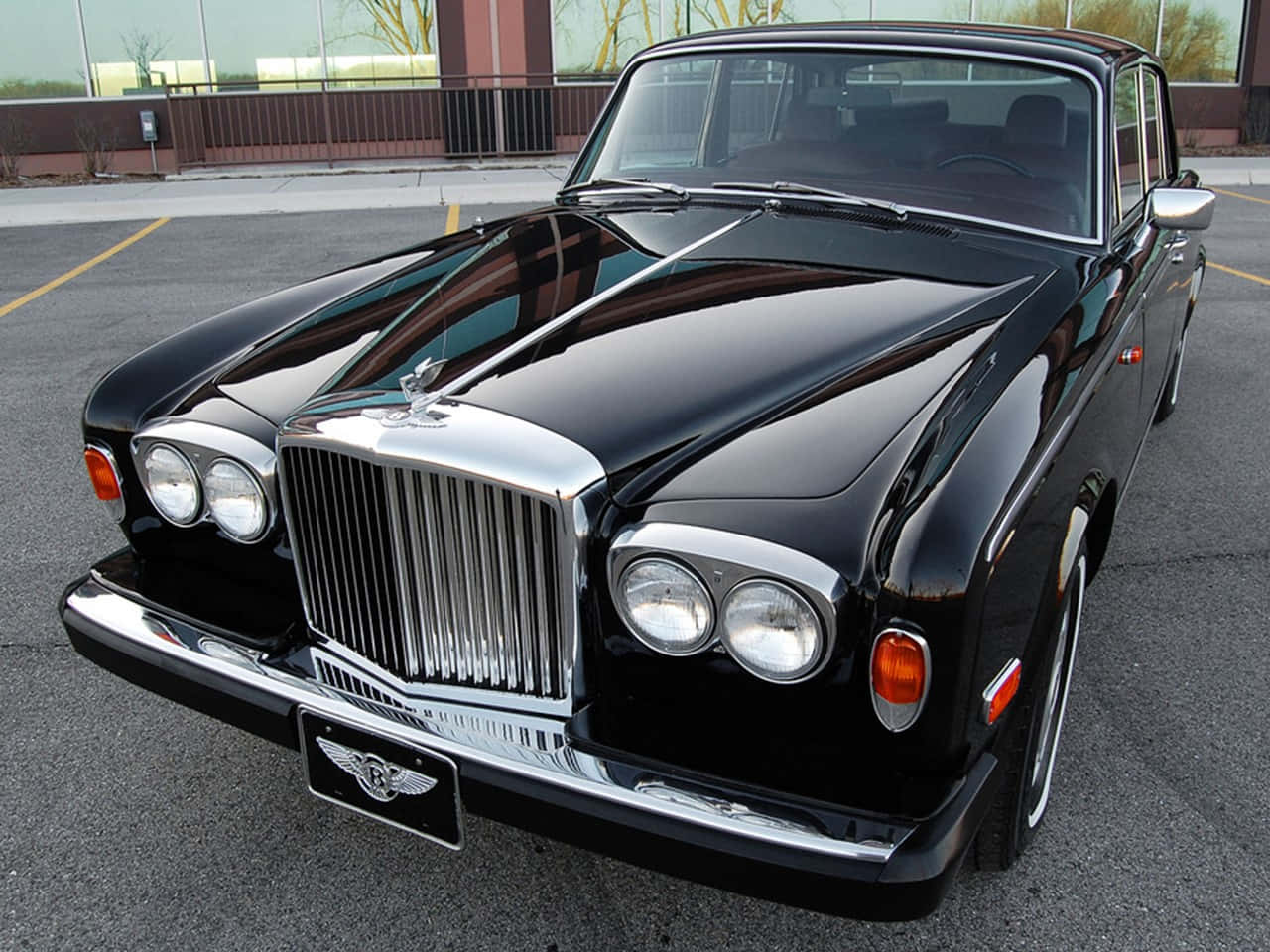 Bentley T2 桌布 壁紙 — 一輛亮黑色賓利 Continental GT 在日光下於鋪砌的停車場中閃耀，背景是現代建築。