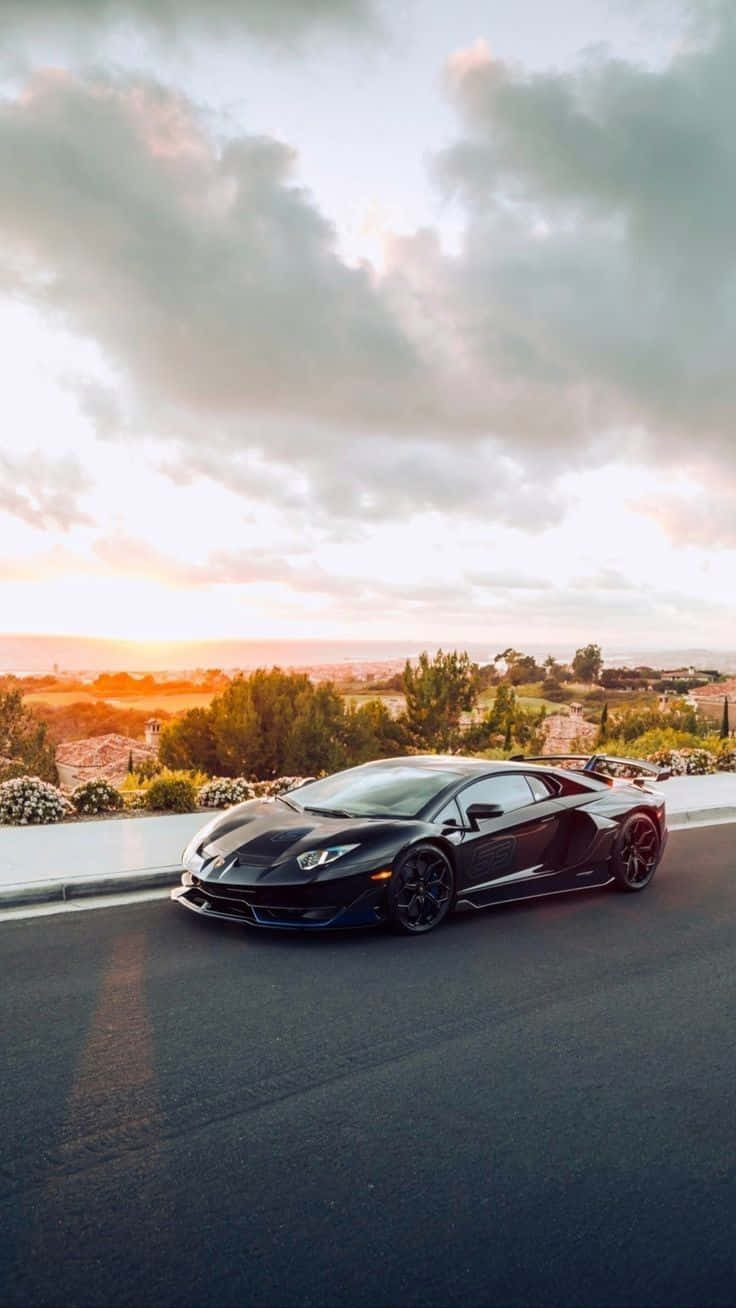 一輛充滿動感的賽車領先群雄 壁紙 — 一架時尚的黑色林寶堅尼 Aventador SVJ 停泊在日落時分的沿海公路上，背景有戲劇性的雲彩和金色光線。