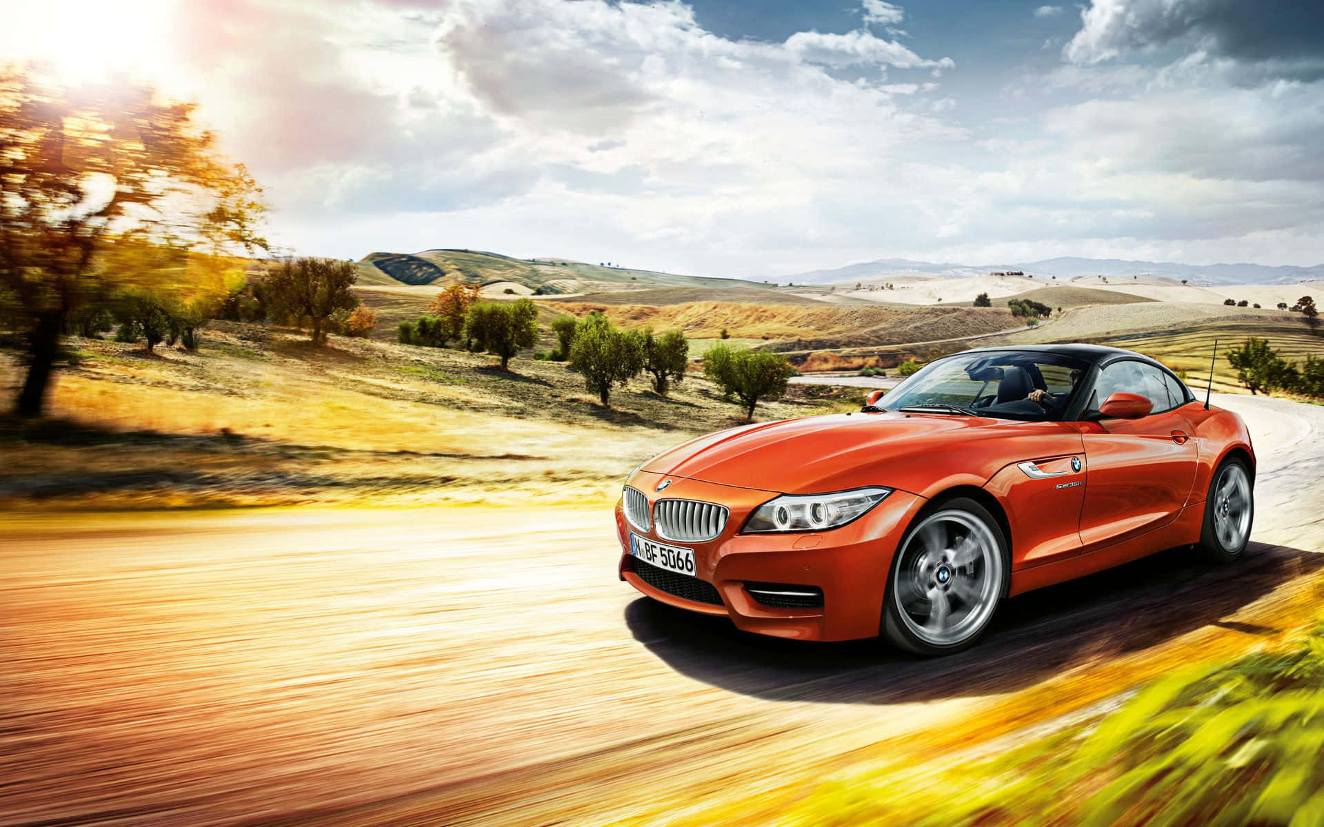 Bmw Z4 桌布 壁紙 — 一輛充滿活力的橙色BMW Z4開篷跑車在部分多雲的天空下，沿著陽光明媚的鄉村道路穿過金色山丘飛馳。