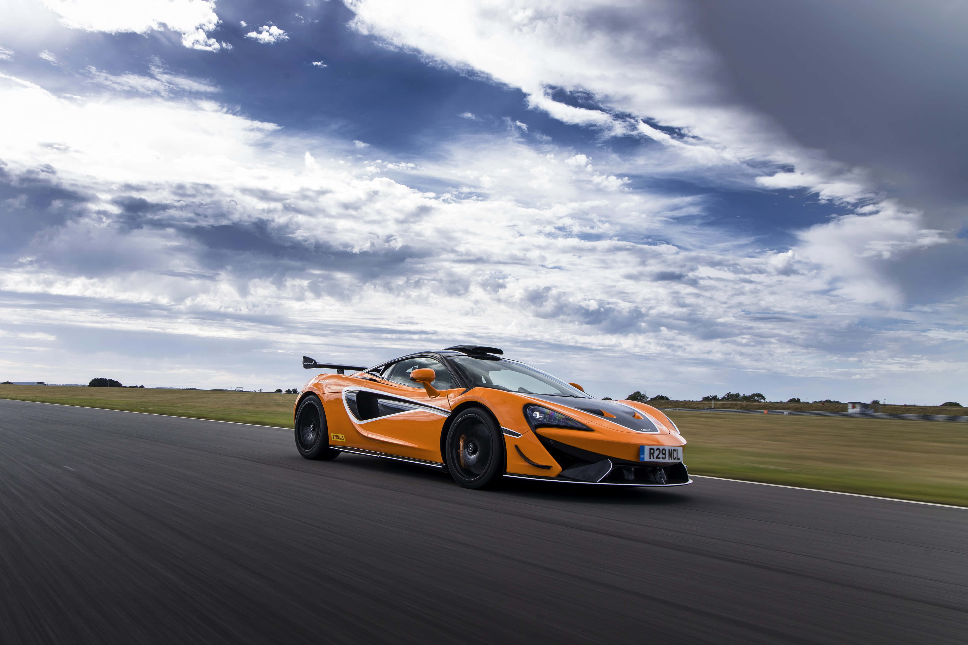 Mclaren 620r 桌布 壁紙 — 一輛充滿活力的橙色McLaren跑車在戲劇性的多雲天空下沿著賽道飛馳。