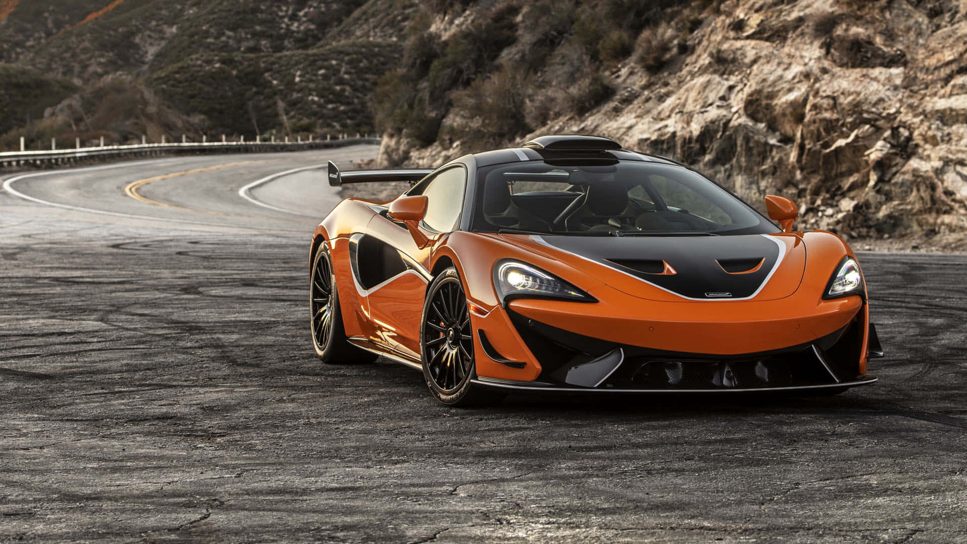 Mclaren 620r 桌布 壁紙 — 一輛充滿活力的橙色McLaren跑車，帶有黑色點綴，在明亮的日光下行駛在蜿蜒的山路上。