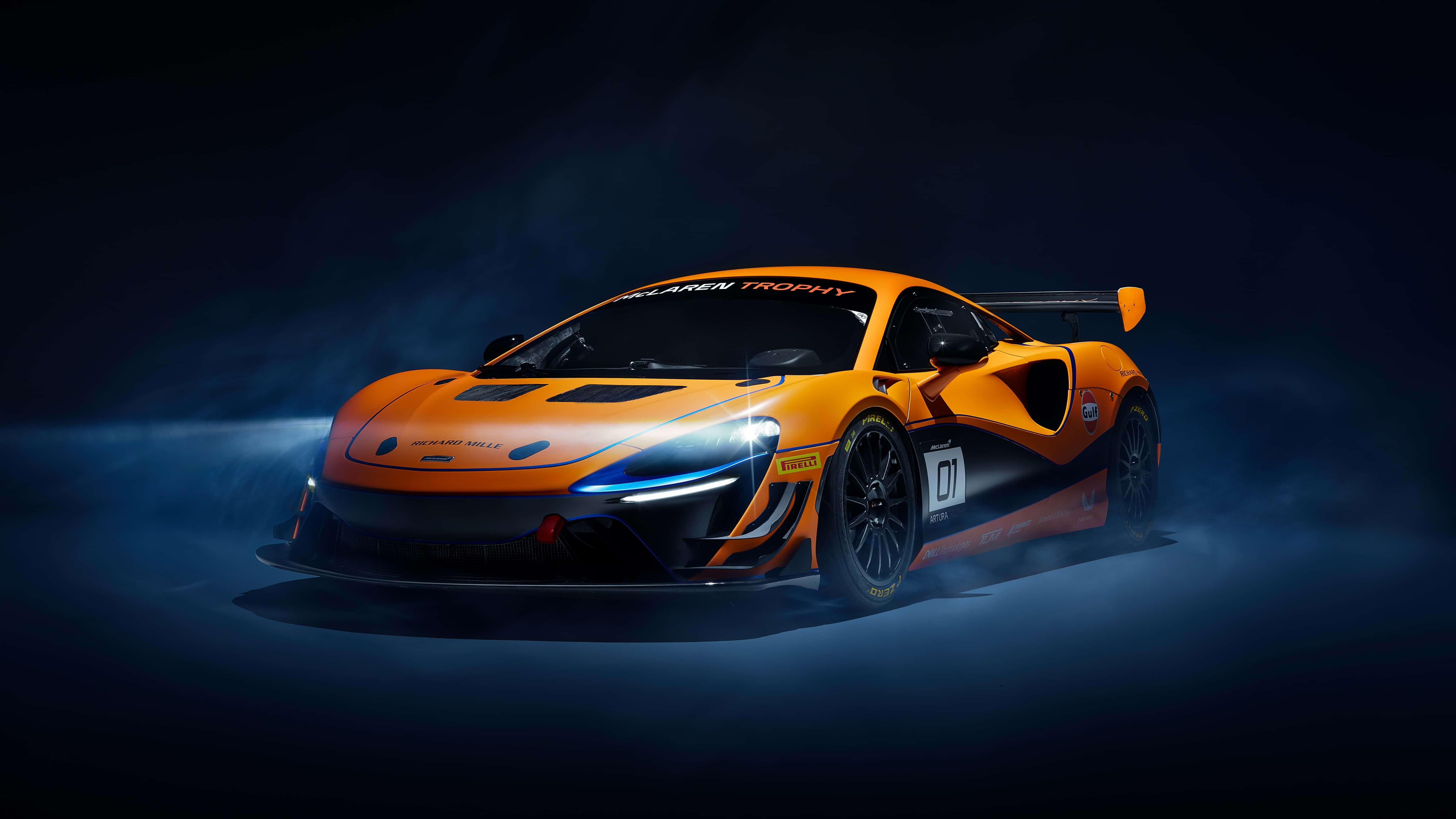 麥拿侖 桌布 壁紙 — 一輛充滿活力的橙色McLaren賽車，在黑暗的攝影棚環境中帶有發光的頭燈和飄散的煙霧。