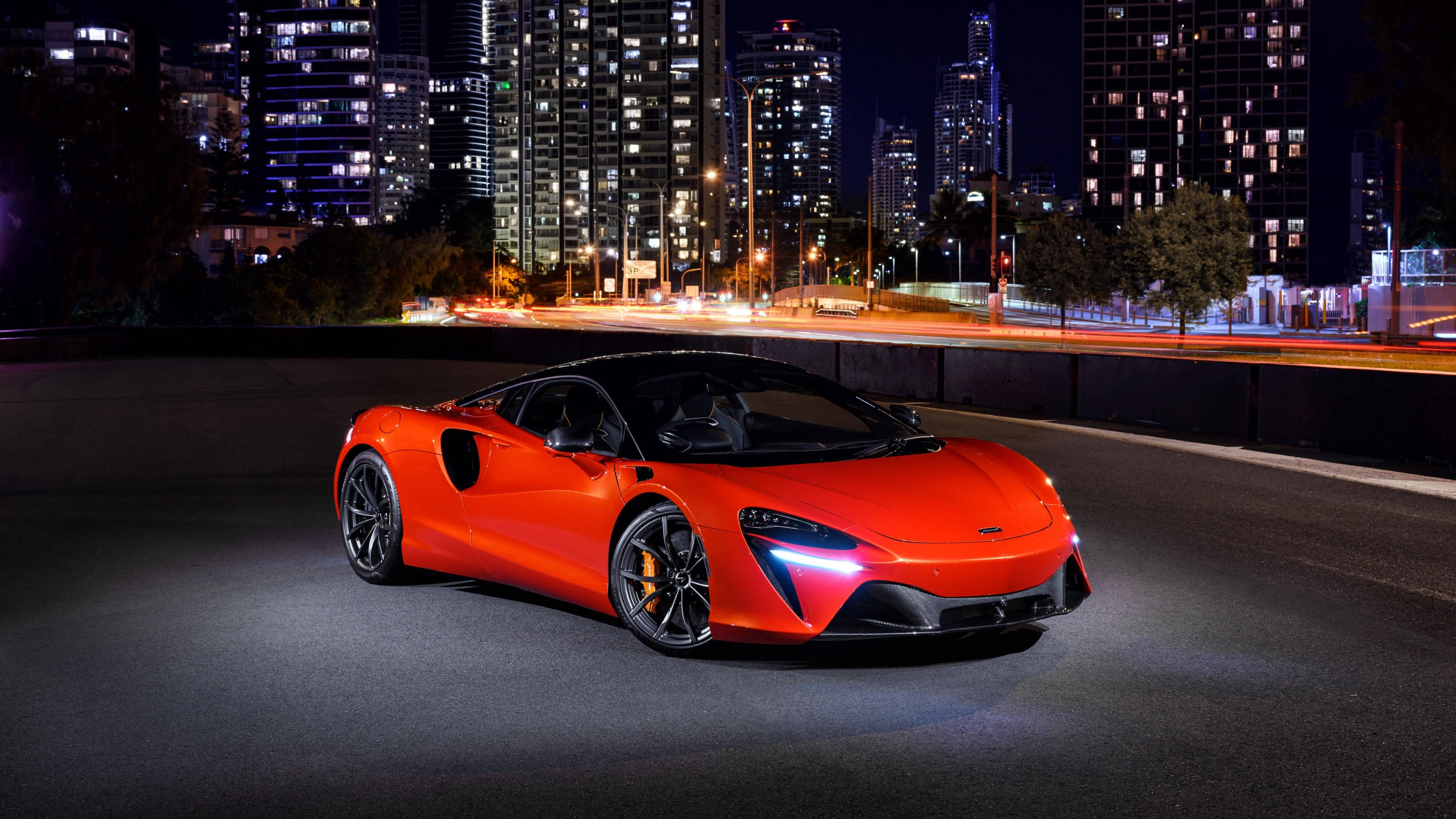 Sports Car 桌布 壁紙 — 一輛充滿活力的橙色McLaren跑車在夜間城市道路上，在街燈下閃耀，背後有模糊的車流痕跡。