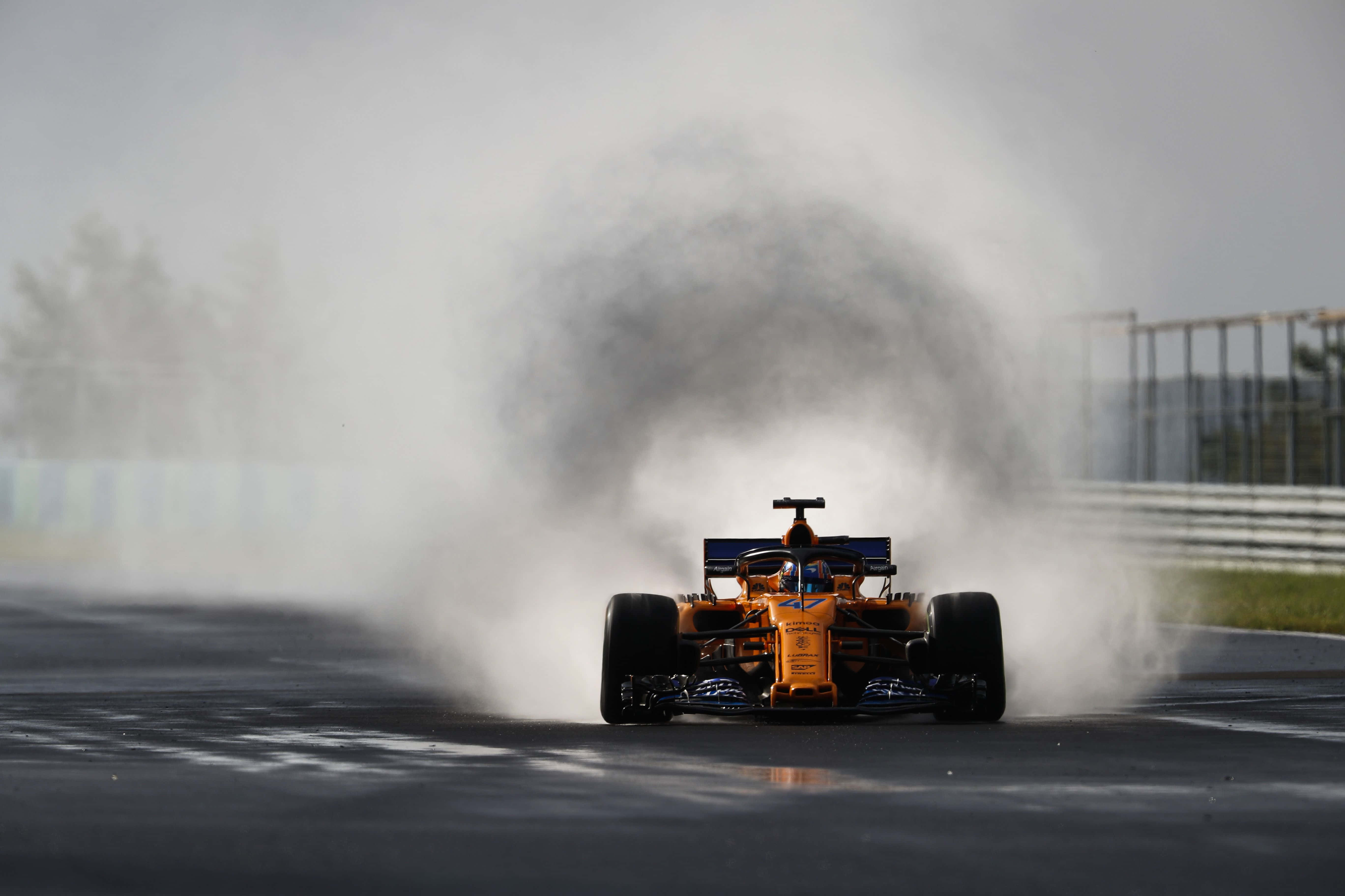 lando norris 桌布 壁紙 — 一輛充滿活力的橙色一級方程式賽車在濕滑彎道中疾馳，在灰色賽道上激起巨大的水霧。