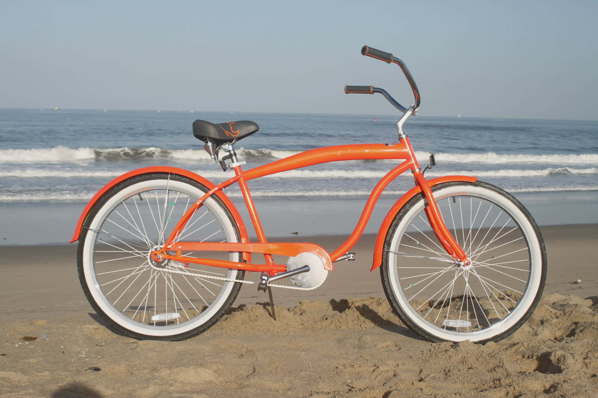 Beach Cruiser 桌布 壁紙 — 一輛充滿活力的橙色沙灘巡洋艦單車停放在沙灘上，海浪在晴朗的天空下翻滾。