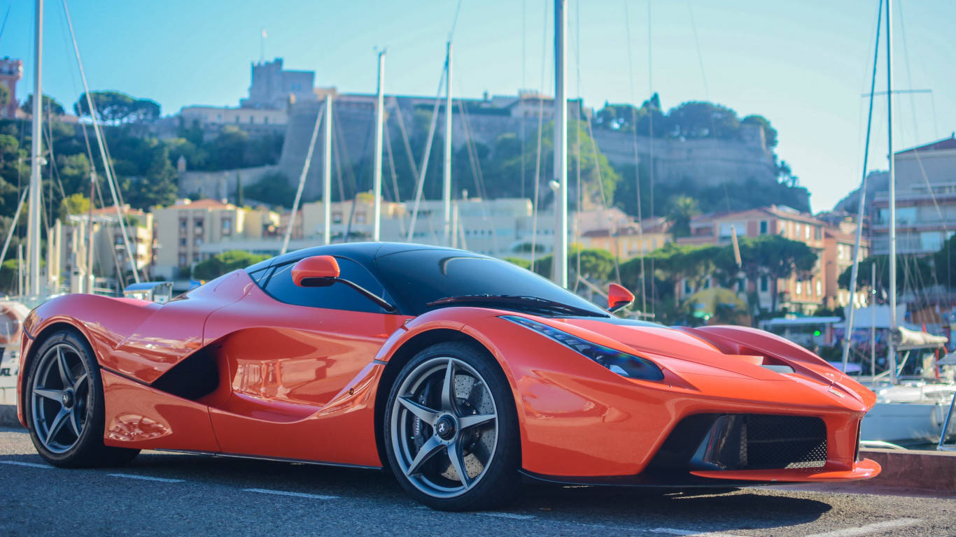 Sports Car 桌布 壁紙 — 一輛充滿活力的橙色法拉利LaFerrari停泊在陽光明媚的碼頭，背景有帆船和一座歷史悠久的山丘堡壘。