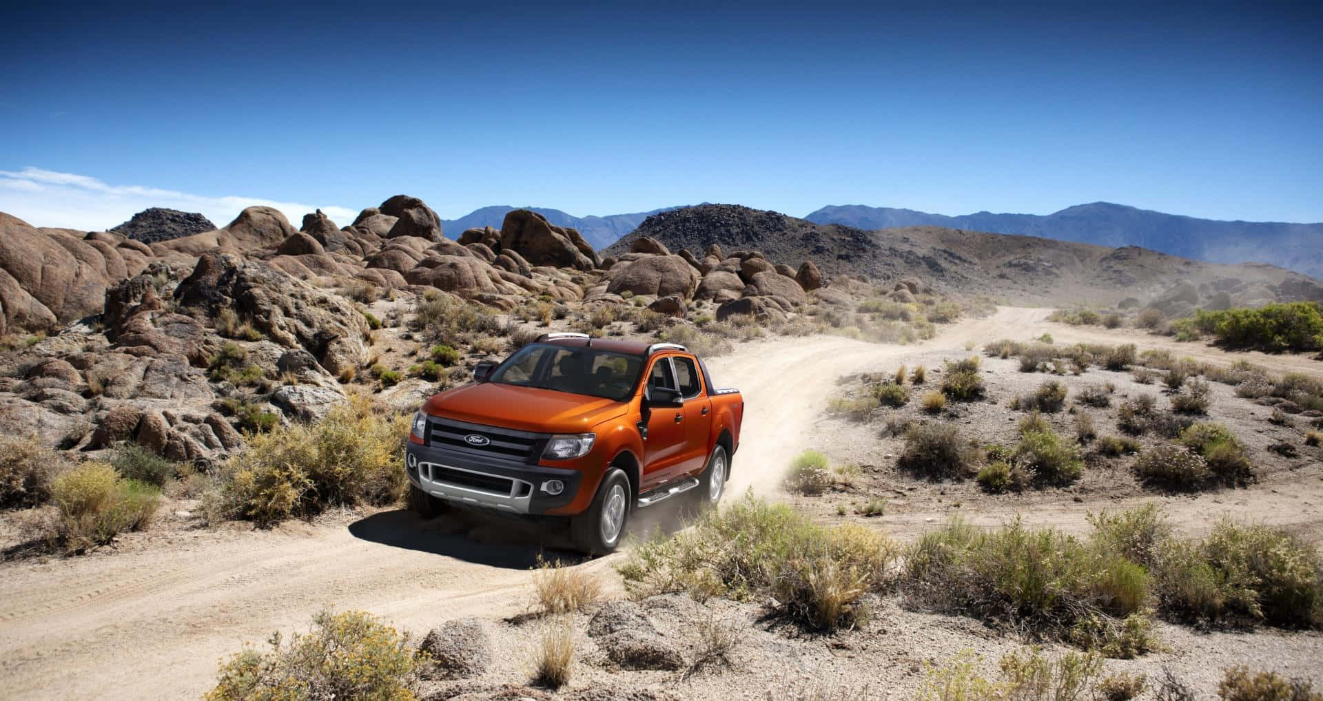 Ford Ranger 桌布 壁紙 — 一輛充滿活力的橙色皮卡車在崎嶇的沙漠小徑上行駛，揚起灰塵，周圍是岩石地形和遠處的山脈，天空清澈湛藍。