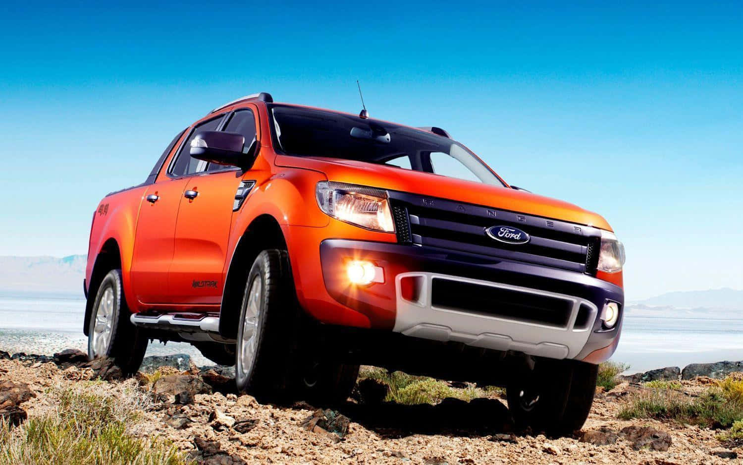 Ford Ranger 桌布 壁紙 — 一輛充滿活力的橙色福特 Ranger 皮卡車，在晴朗的藍天下，停泊在岩石海岸線上，遠處有山巒。