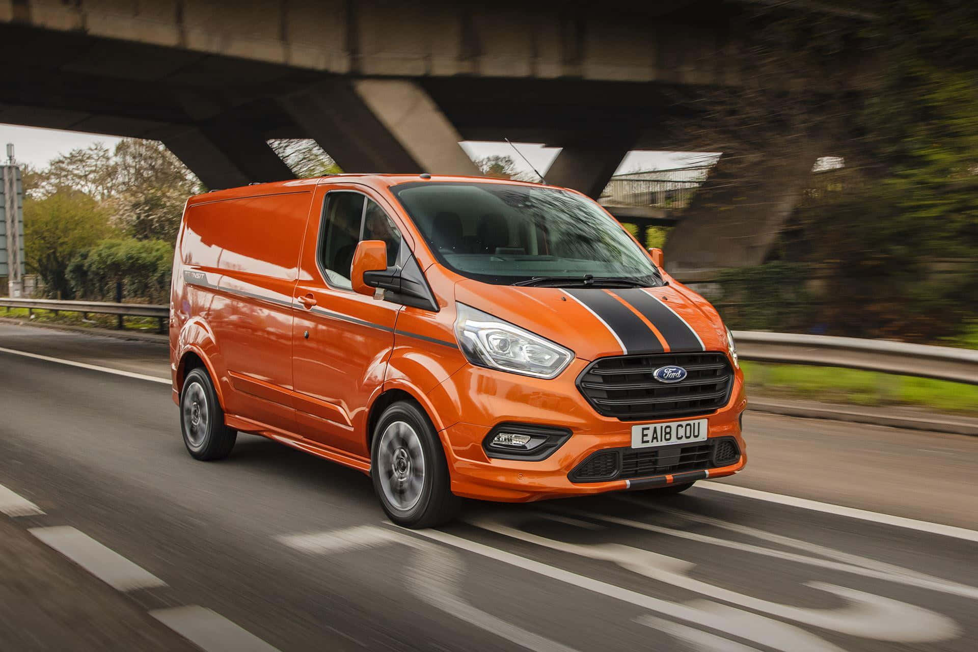 Ford Transit 桌布 壁紙 — 一輛充滿活力的橙色福特全順客貨車帶有黑色賽車條紋，在綠樹成蔭的道路上飛馳於混凝土高架橋下。