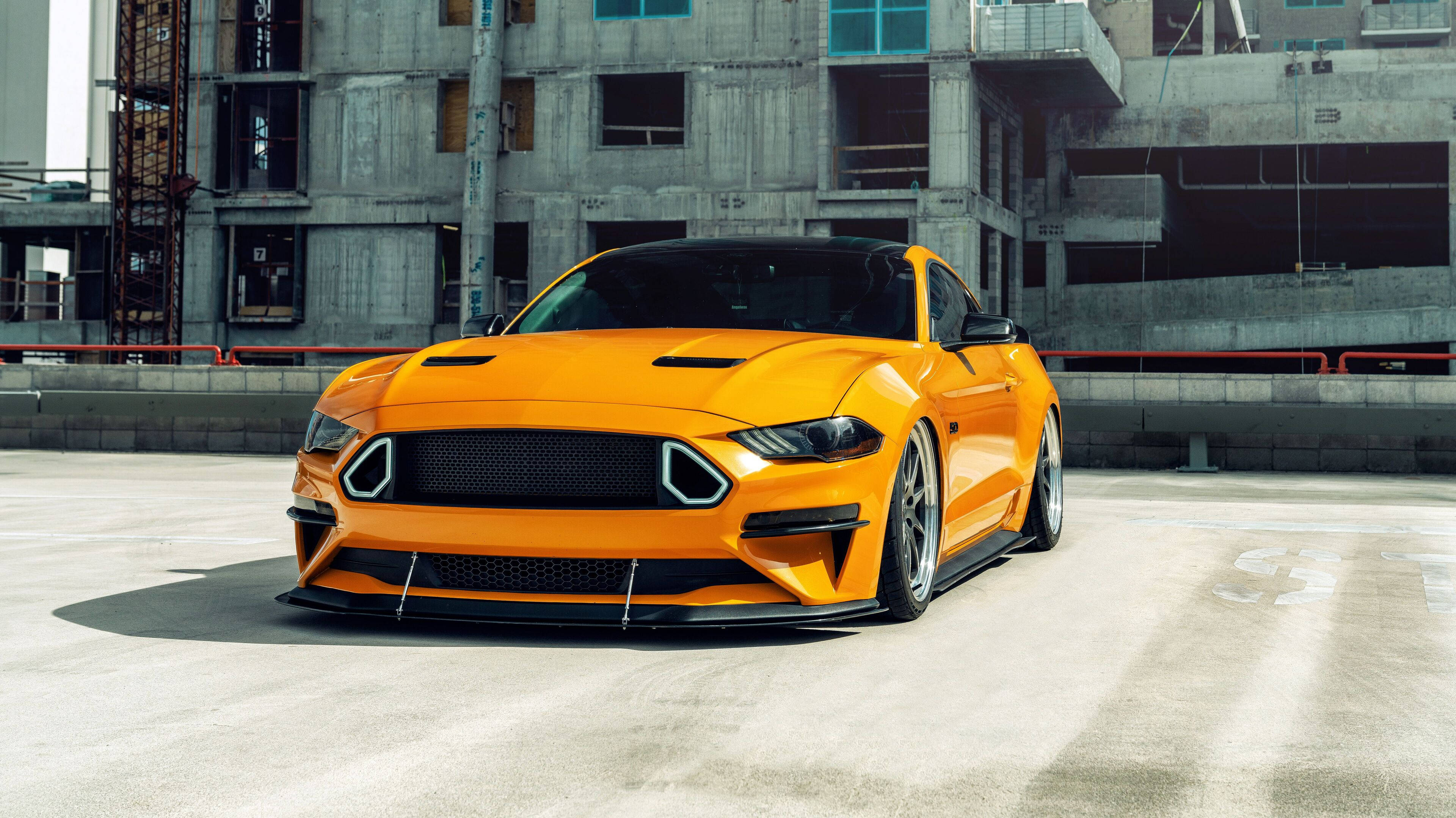 mustang HD 桌布 壁紙 — 一輛充滿活力的橙色福特野馬，具有侵略性的空氣動力學設計，停在明亮的日光下的混凝土建築工地前。