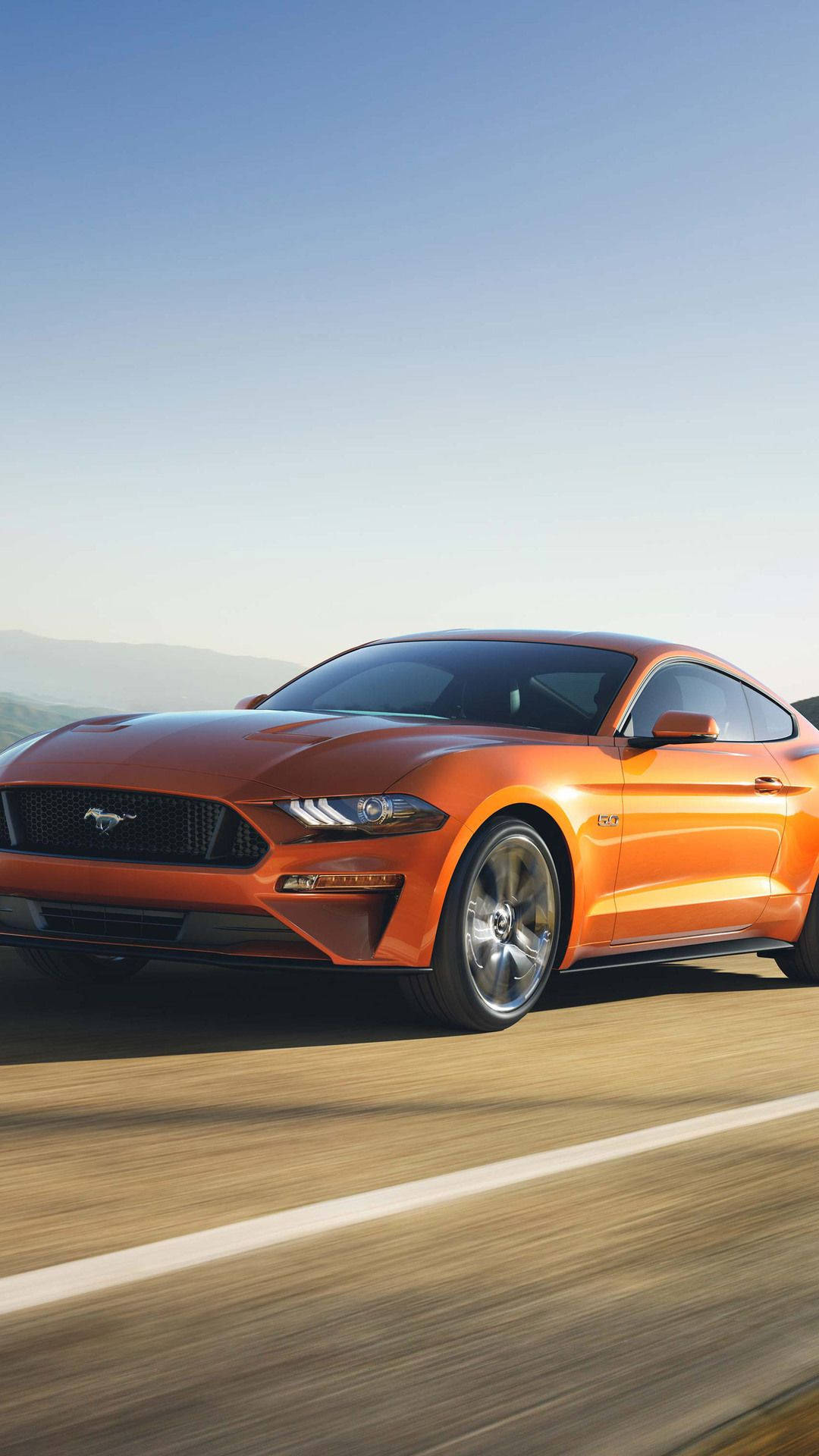 Ford 桌布 壁紙 — 一輛充滿活力的橙色福特野馬在陽光明媚的高速公路上疾馳，背景是模糊的動態和遠處的山丘，天空清澈湛藍。