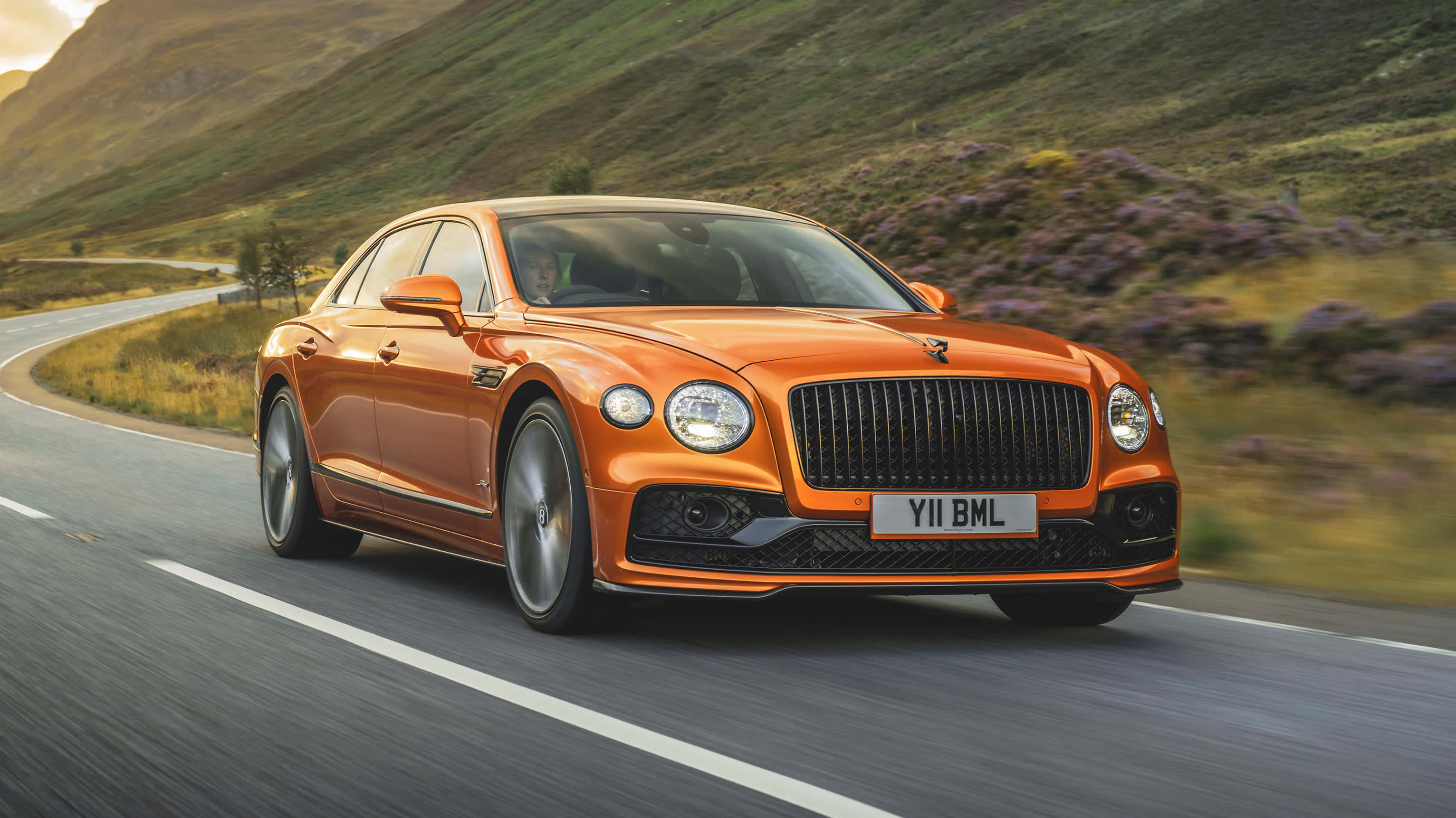 Bentley Flying Spur 桌布 壁紙 — 一輛充滿活力的橙色賓利飛馳在蜿蜒的山路上，周圍是紫色的石楠花和綠色的山丘，正值黃金時刻。