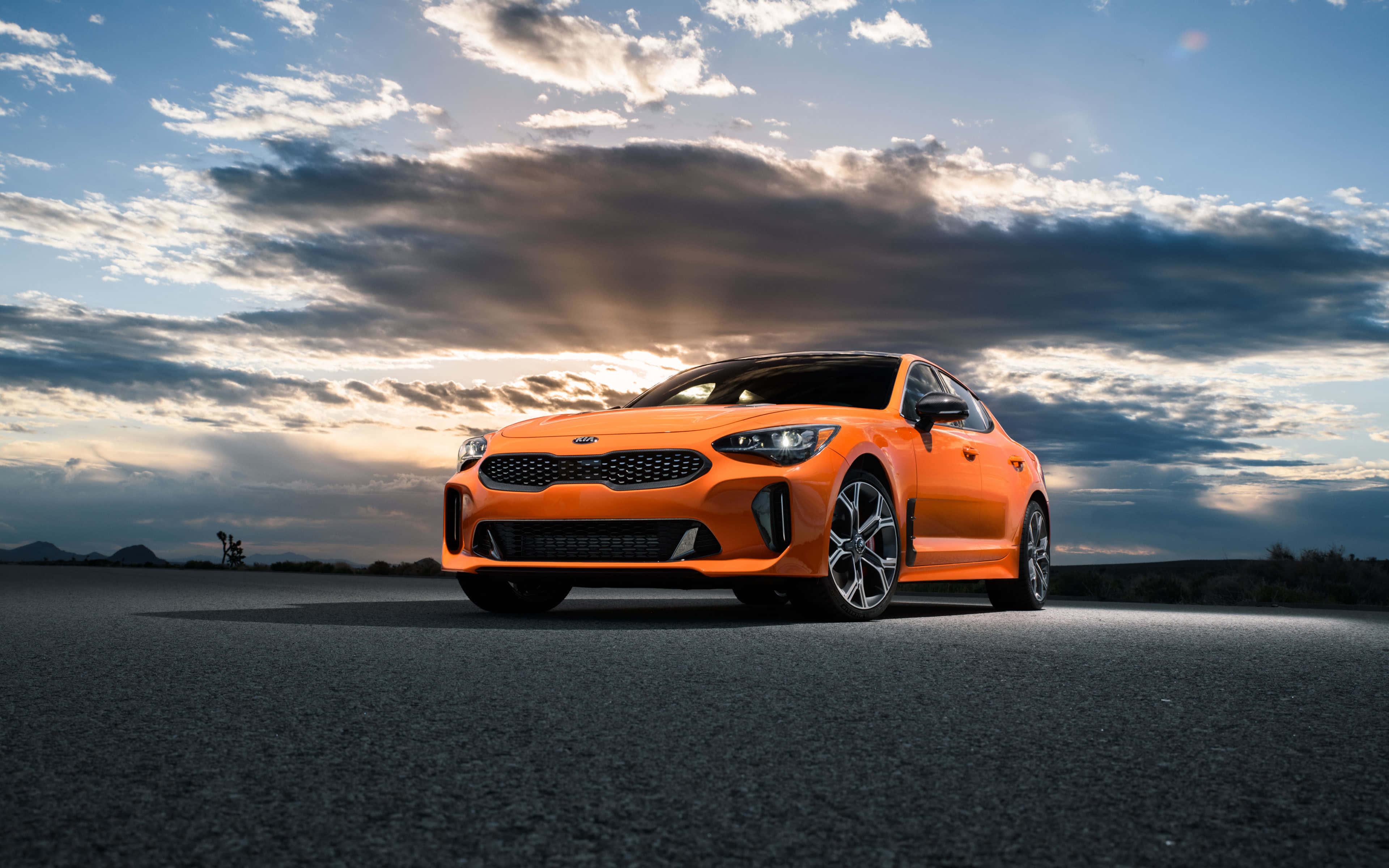Kia Stinger 桌布 壁紙 — 一輛充滿活力的橙色跑車在戲劇性的日落雲彩下，在開闊的瀝青路上閃閃發光。