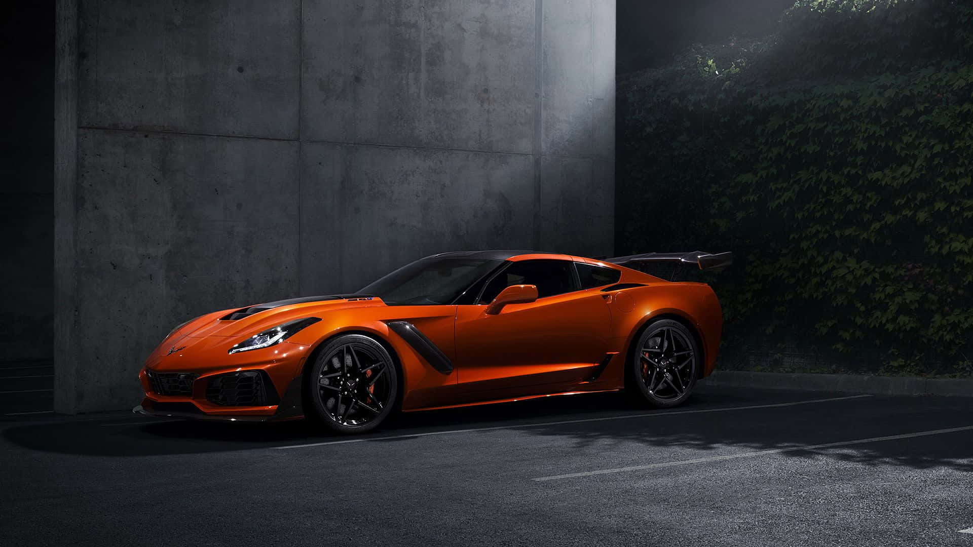 corvette 桌布 壁紙 — 一輛充滿活力的橙色跑車帶有黑色點綴，在戲劇性的聚光燈下，以混凝土牆和常春藤覆蓋的背景為襯托，閃閃發光。