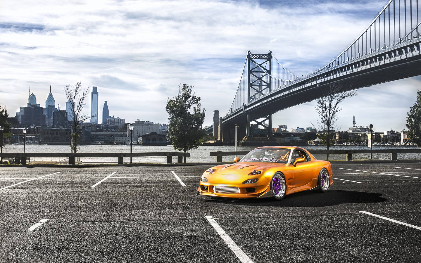 Mazda 桌布 壁紙 — 一輛充滿活力的橙色跑車配紫色輪圈，停在空地上，背景是陰天、橋樑和城市天際線。
