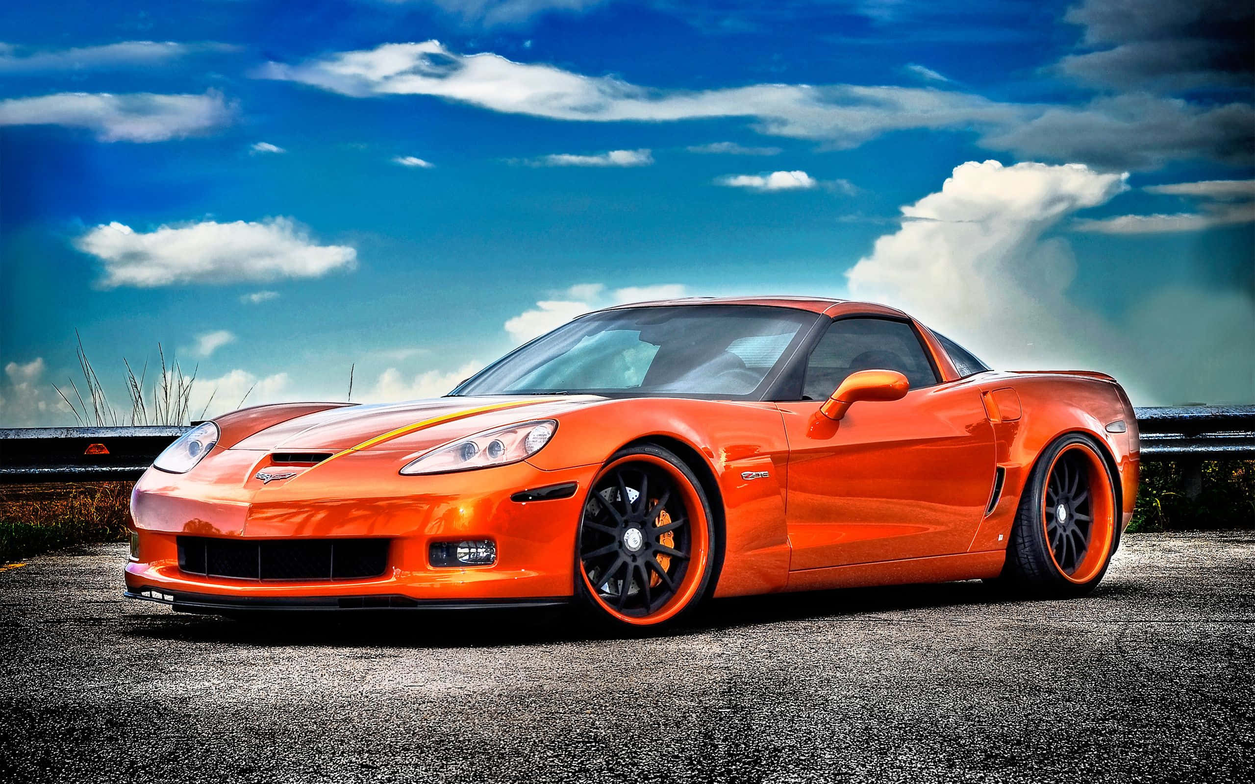 Chevrolet Corvette Z06 桌布 壁紙 — 一輛充滿活力的橙色跑車配黑色輪圈，停在路邊，背景是戲劇性的藍天和蓬鬆的白雲。