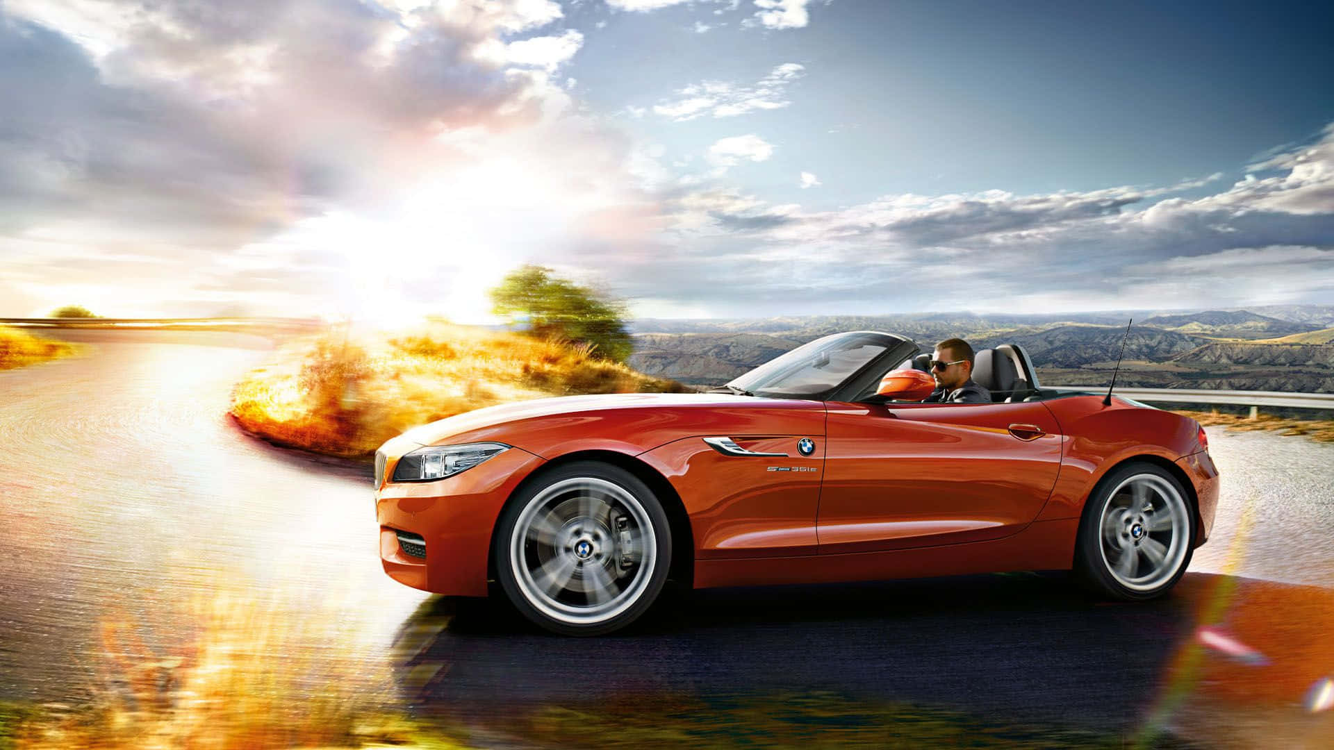Bmw Z4 桌布 壁紙 — 一輛充滿活力的橙色開篷跑車在日落時分沿著蜿蜒的道路疾馳，司機享受著露天駕駛的樂趣。