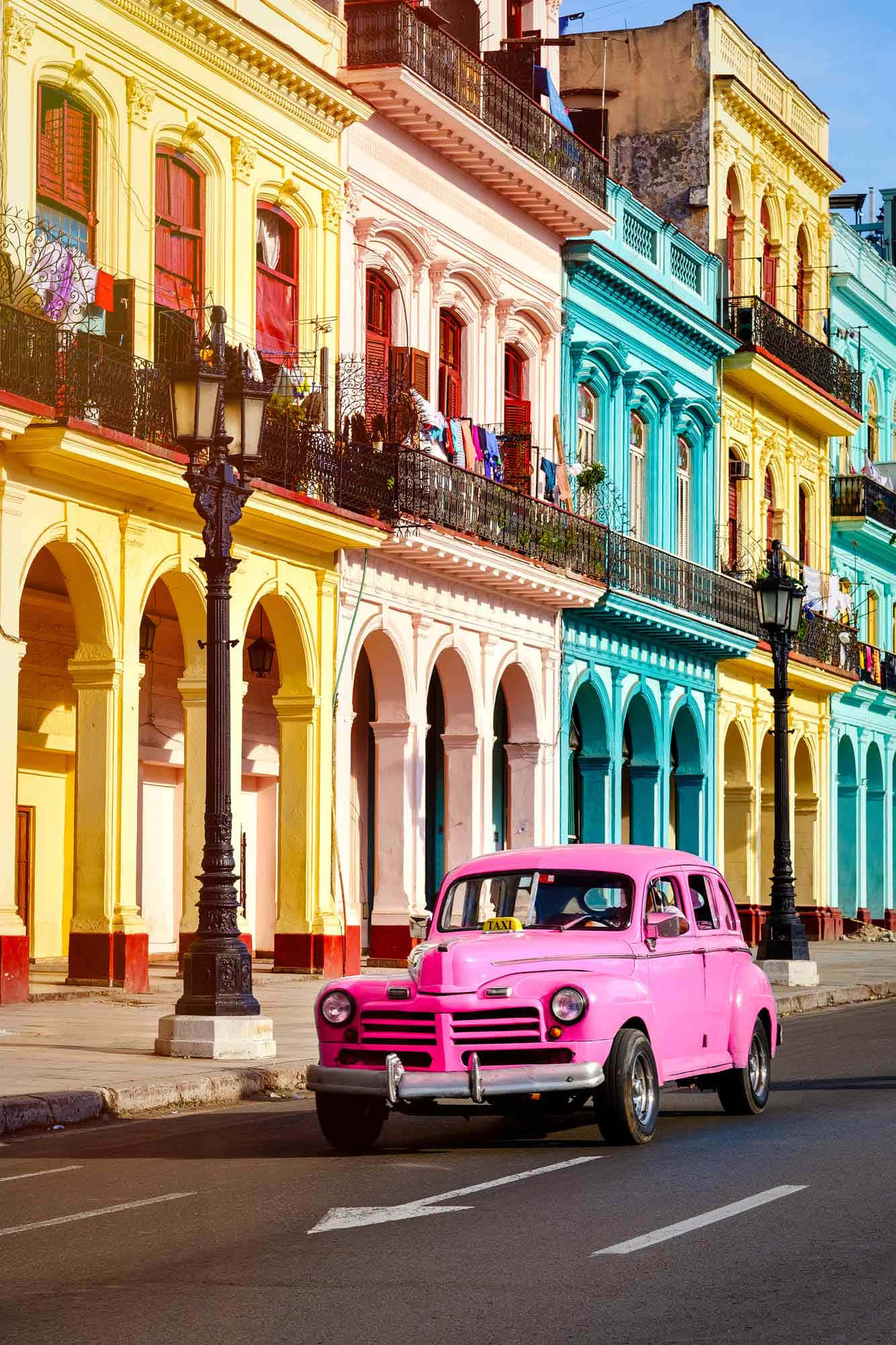 Cuba 桌布 壁紙 — 一輛充滿活力的粉紅色復古汽車在古巴一條陽光普照、兩旁色彩繽紛殖民建築的街道上行駛。