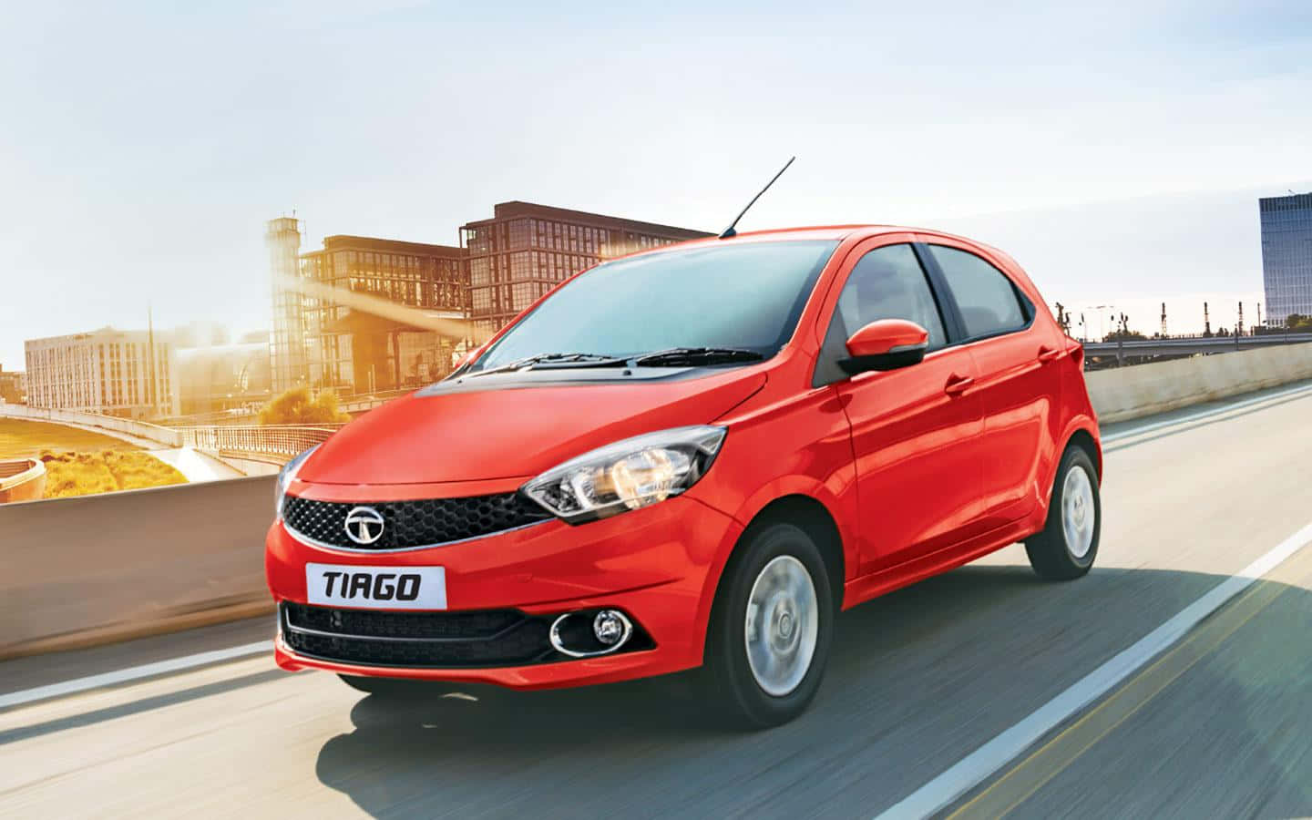 Tata Motors 桌布 壁紙 — 一輛充滿活力的紅色Tata Tiago掀背車在陽光明媚的城市高速公路上飛馳，背景是現代建築。