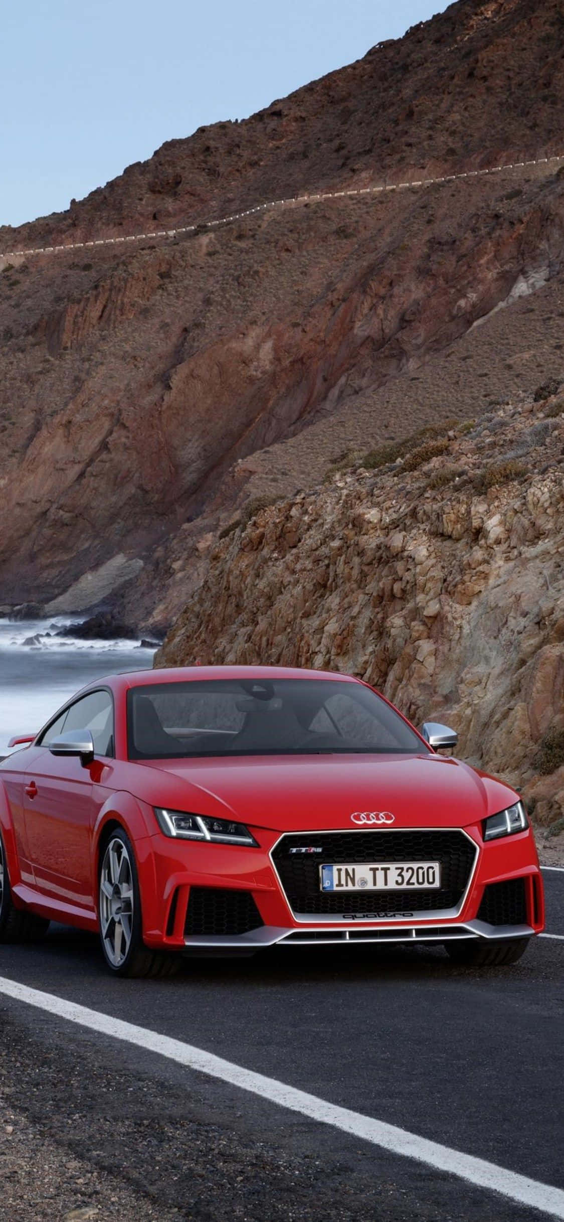 Audi Tt 桌布 壁紙 — 一輛充滿活力的紅色奧迪TT RS跑車沿著海岸懸崖道路行駛，背景是崎嶇的棕色山脈和拍打的海浪。