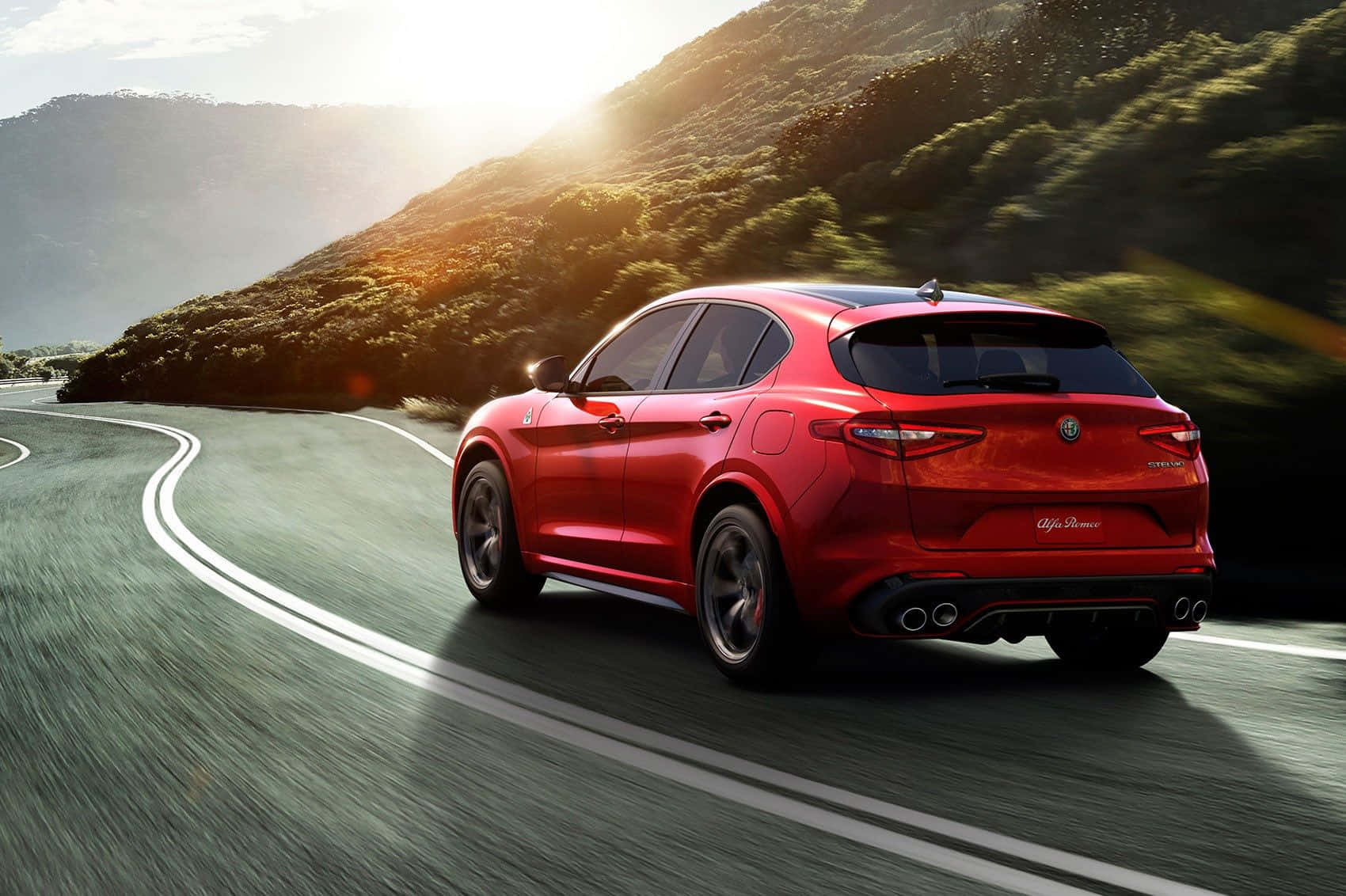 Alfa Romeo Stelvio 桌布 壁紙 — 一輛充滿活力的紅色愛快羅密歐SUV在蜿蜒的山路上疾馳，沐浴在金色的夕陽光中。