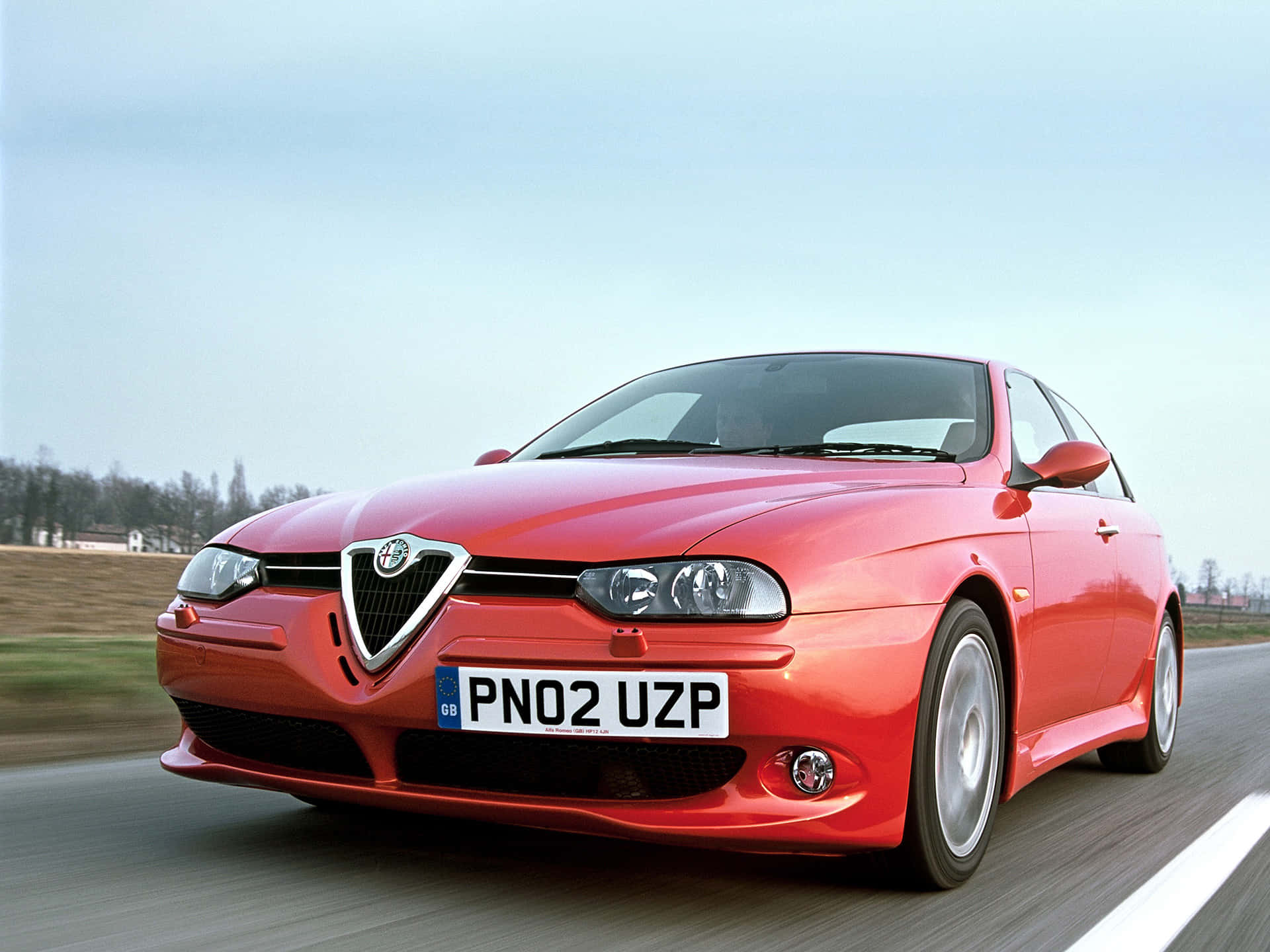 Alfa Romeo 156 桌布 壁紙 — 一輛充滿活力的紅色愛快羅密歐跑車在晴朗的天空下，以模糊的動態沿著鄉村道路疾馳。