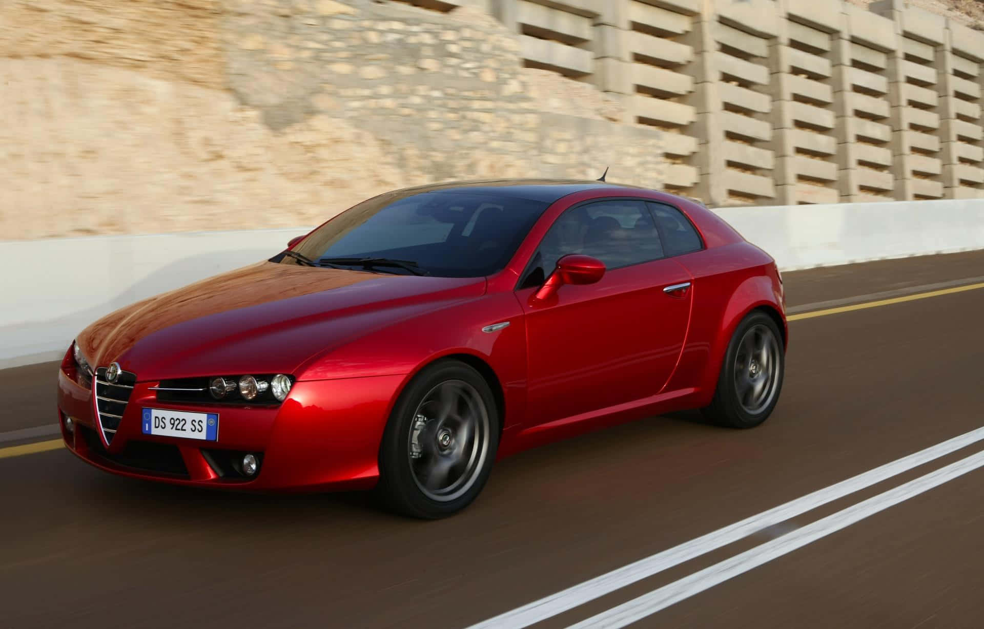 Alfa Romeo Brera 桌布 壁紙 — 一輛充滿活力的紅色愛快羅密歐跑車，在陽光明媚的道路上疾馳，石牆和白色護欄呈現動態模糊效果。