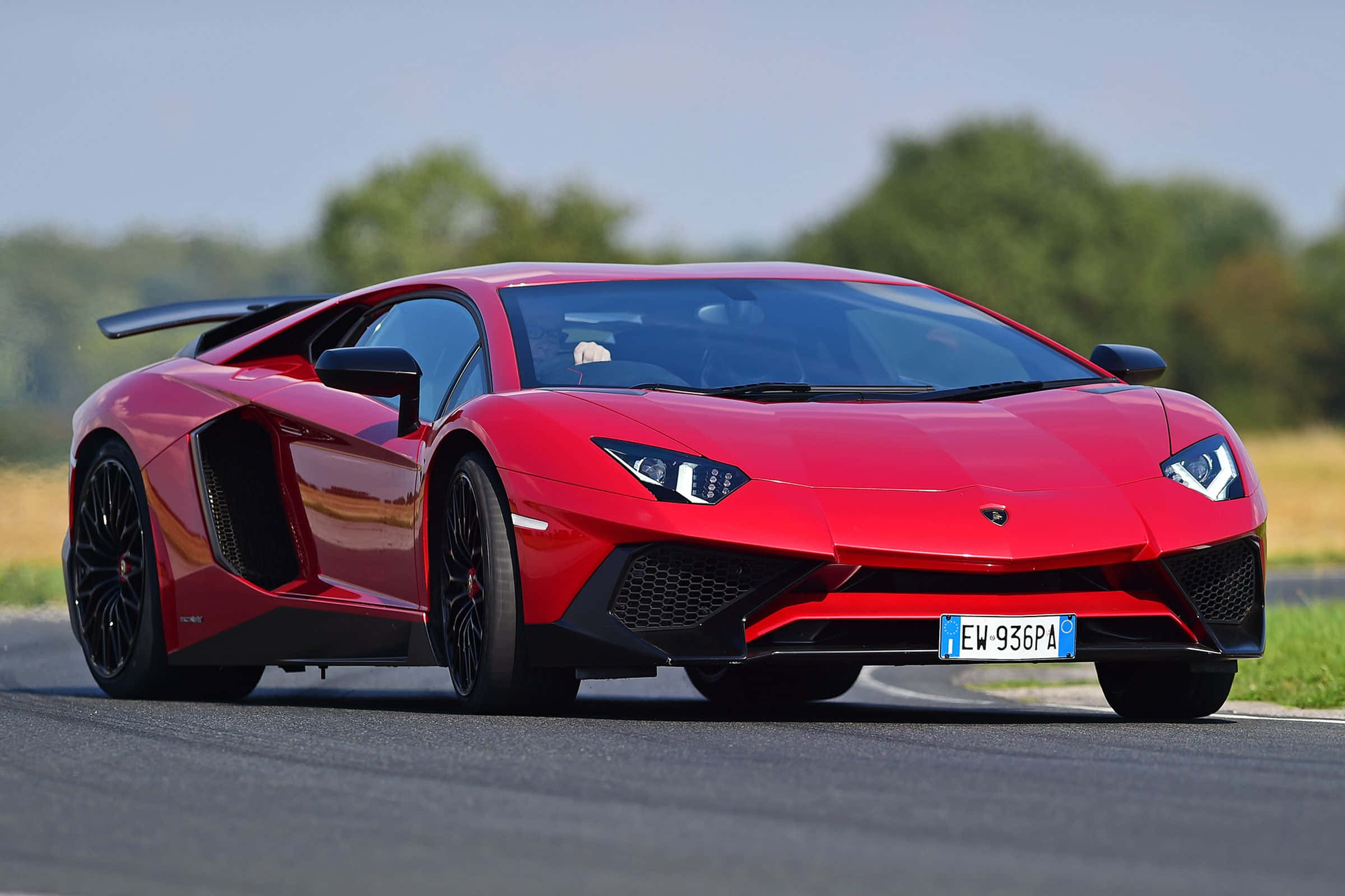 cars 桌布 壁紙 — 一輛充滿活力的紅色林寶堅尼Aventador跑車，帶有黑色飾邊，在晴朗的天空下沿著一條兩旁綠樹成蔭的瀝青道路飛馳。