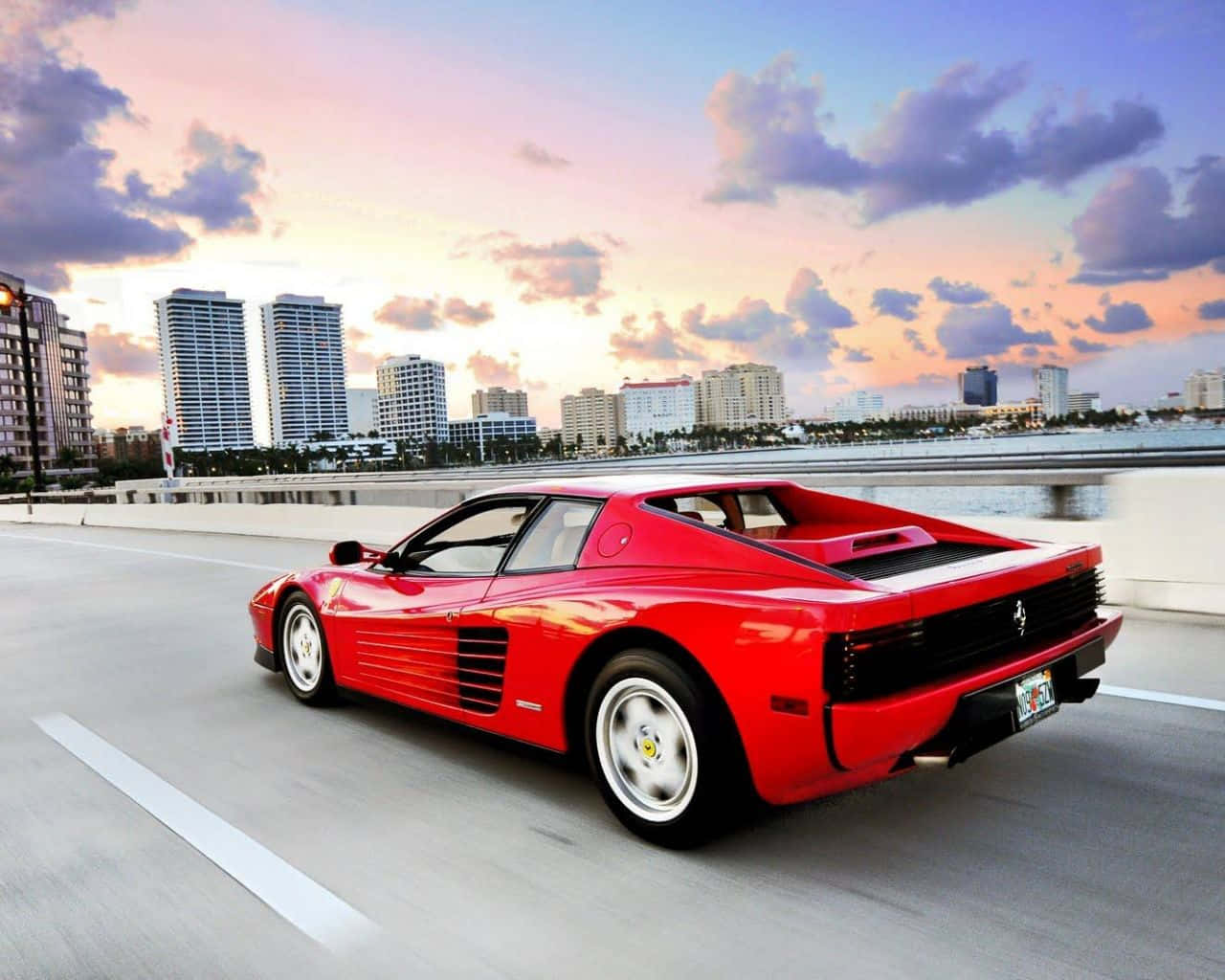 Ferrari Testarossa 桌布 壁紙 — 一輛充滿活力的紅色法拉利F40在日落時分於城市橋樑上飛馳，背景有摩天大樓和雲朵。