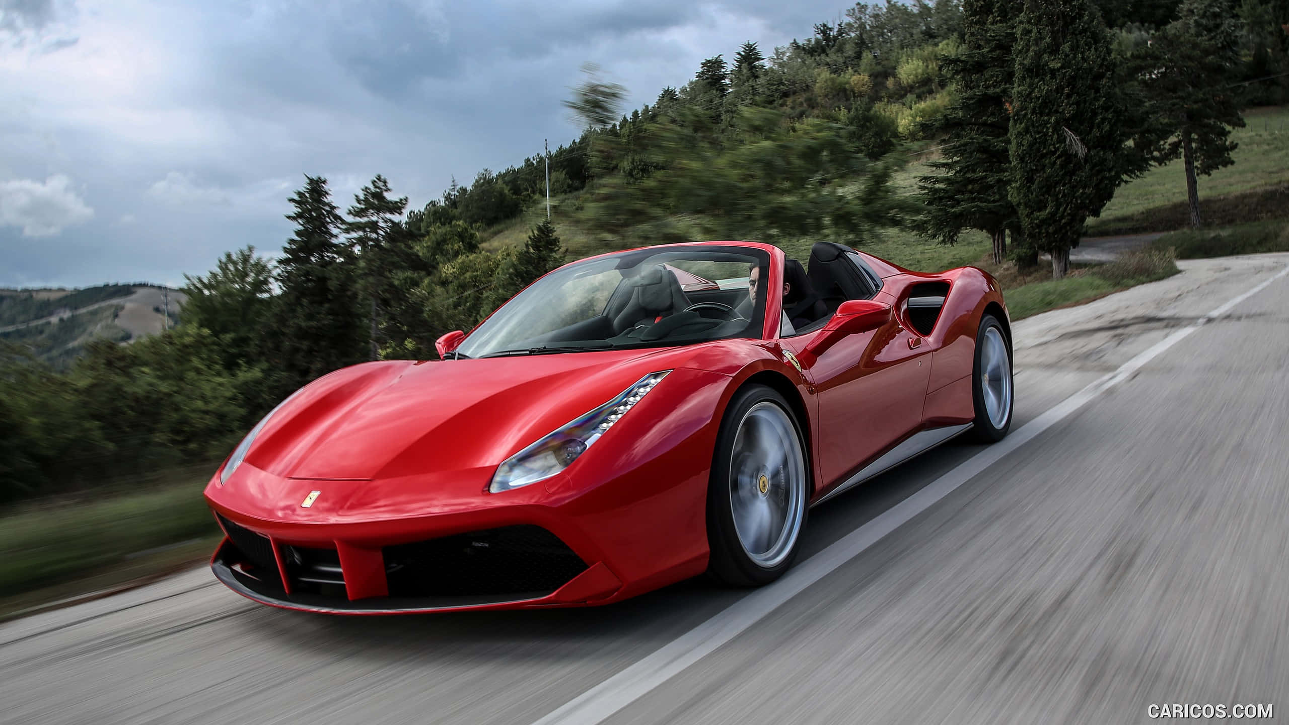 Ferrari 488 Spider 桌布 壁紙 — 一輛充滿活力的紅色法拉利跑車在蜿蜒的山路上疾馳，背景是模糊的綠樹和多雲的天空。