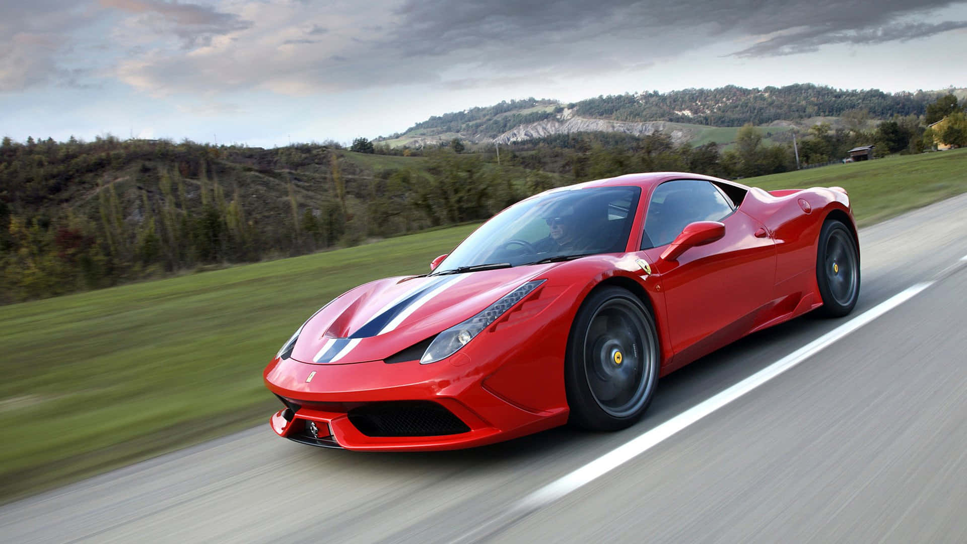 Ferrari 458 Speciale 桌布 壁紙 — 一輛充滿活力的紅色法拉利跑車沿著蜿蜒的鄉村道路飛馳，背景是模糊的綠色山丘和多雲的天空。